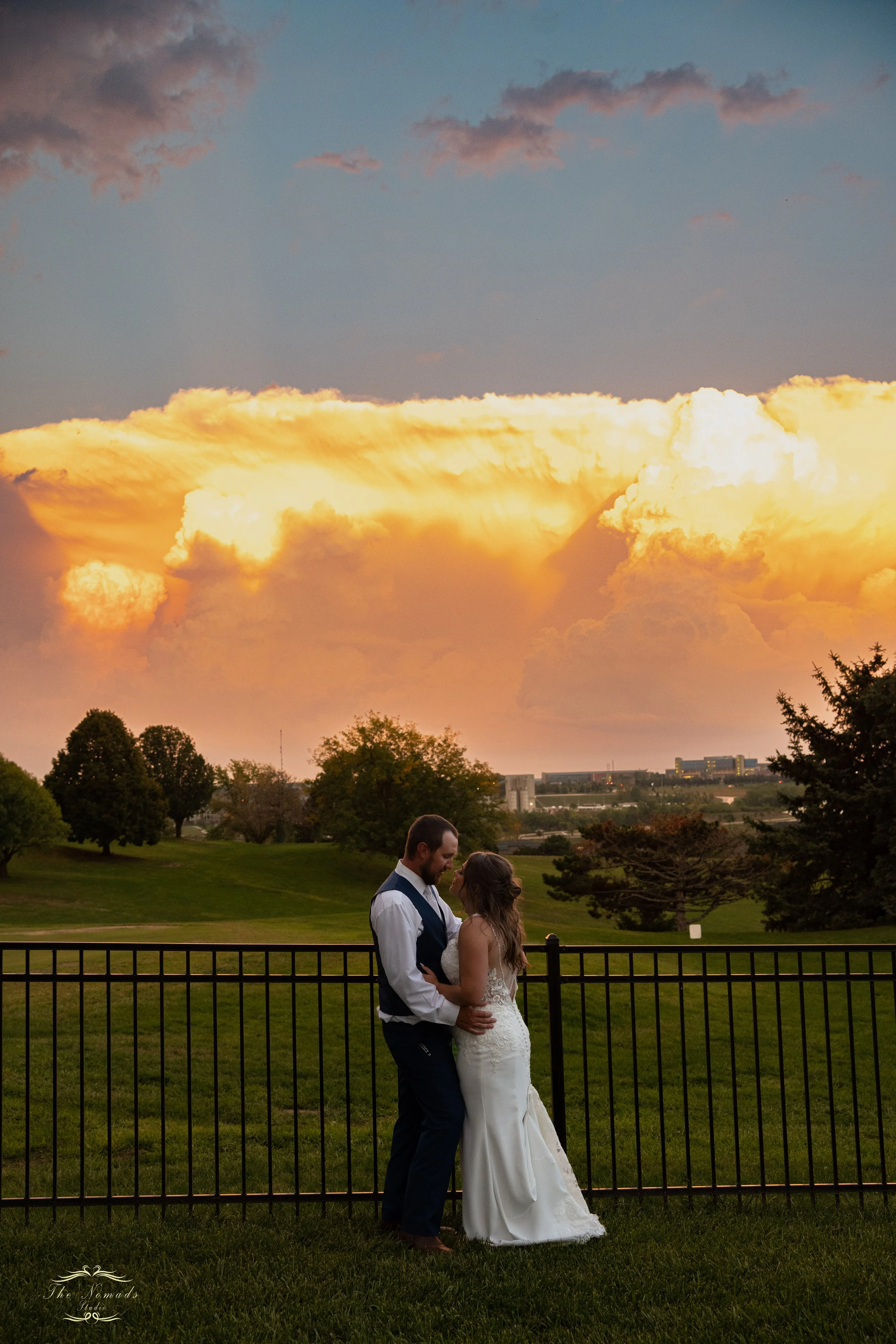 A bride and groom standing close together behind a fence in a park, with trees and a cityscape in the background during sunset, under a sky with large yellow and gray clouds.
