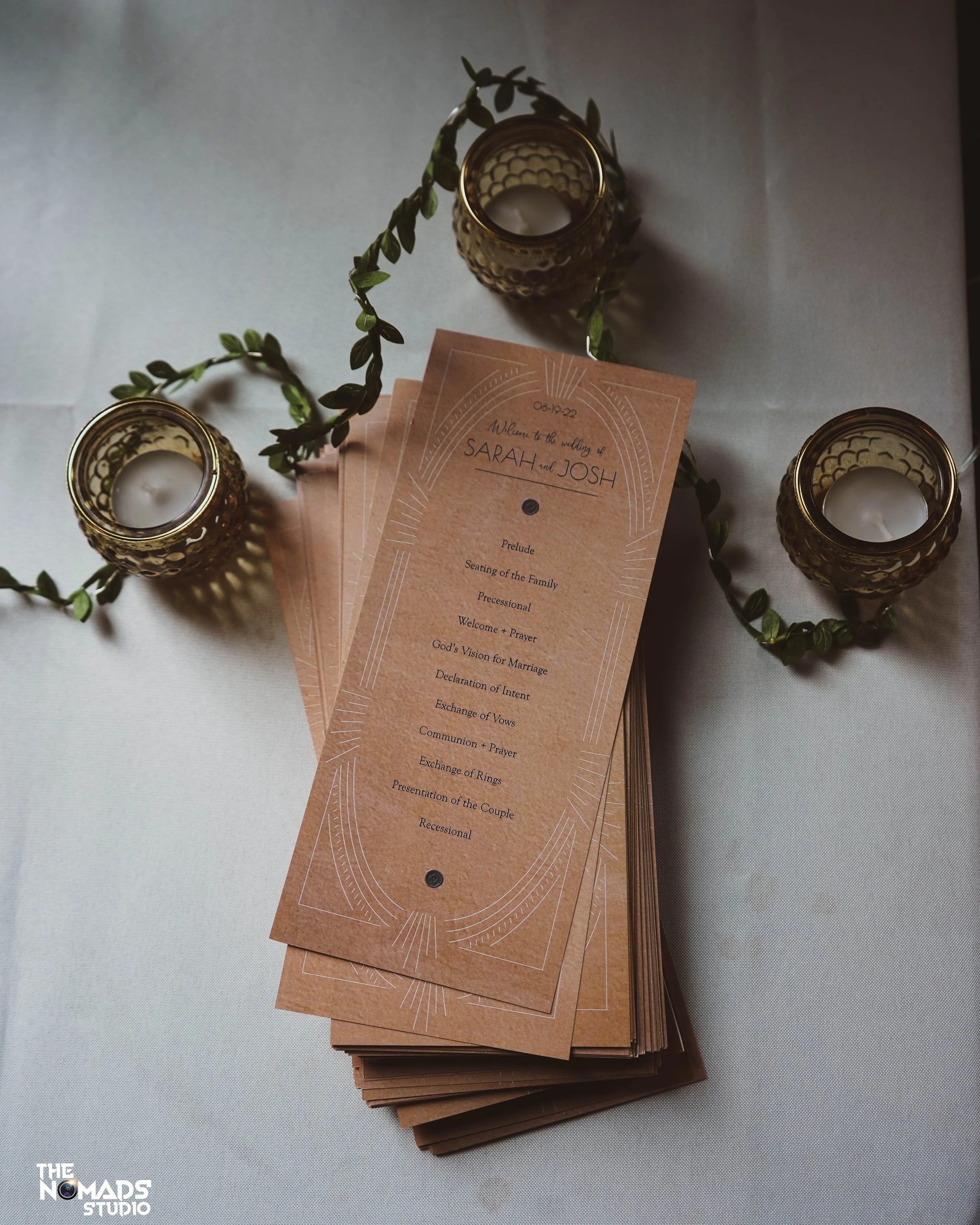 A wedding program brochure, candles in decorative holders, and greenery on a white tablecloth.