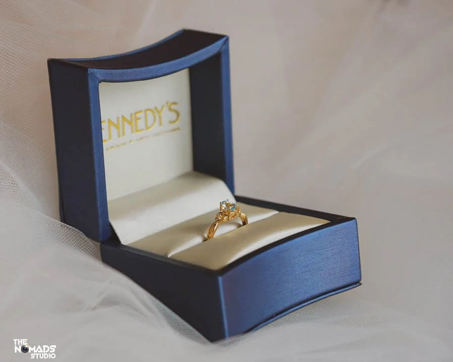 A gold ring with a gemstone in a blue and white jewelry box with "Kennedy's" branding inside.