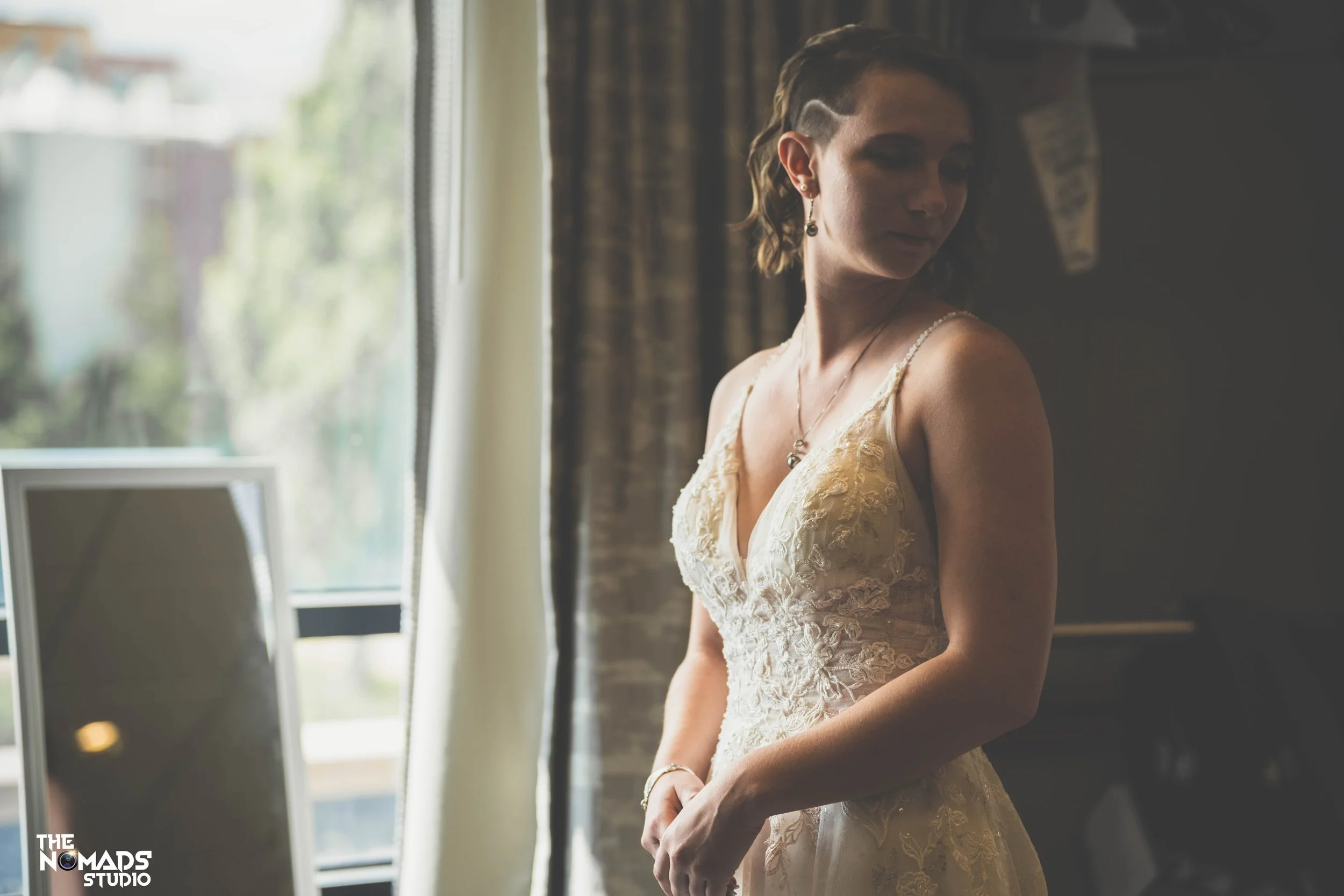 A woman with short hair wearing a lace wedding dress standing near a window, looking down with a soft expression.