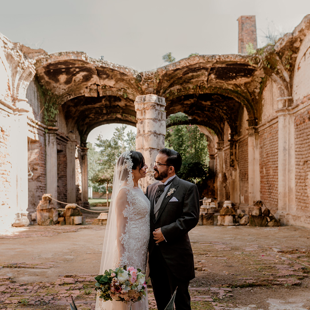 Bride and groom standing close together in wedding attire, holding a bouquet, with ancient ruins in the background.