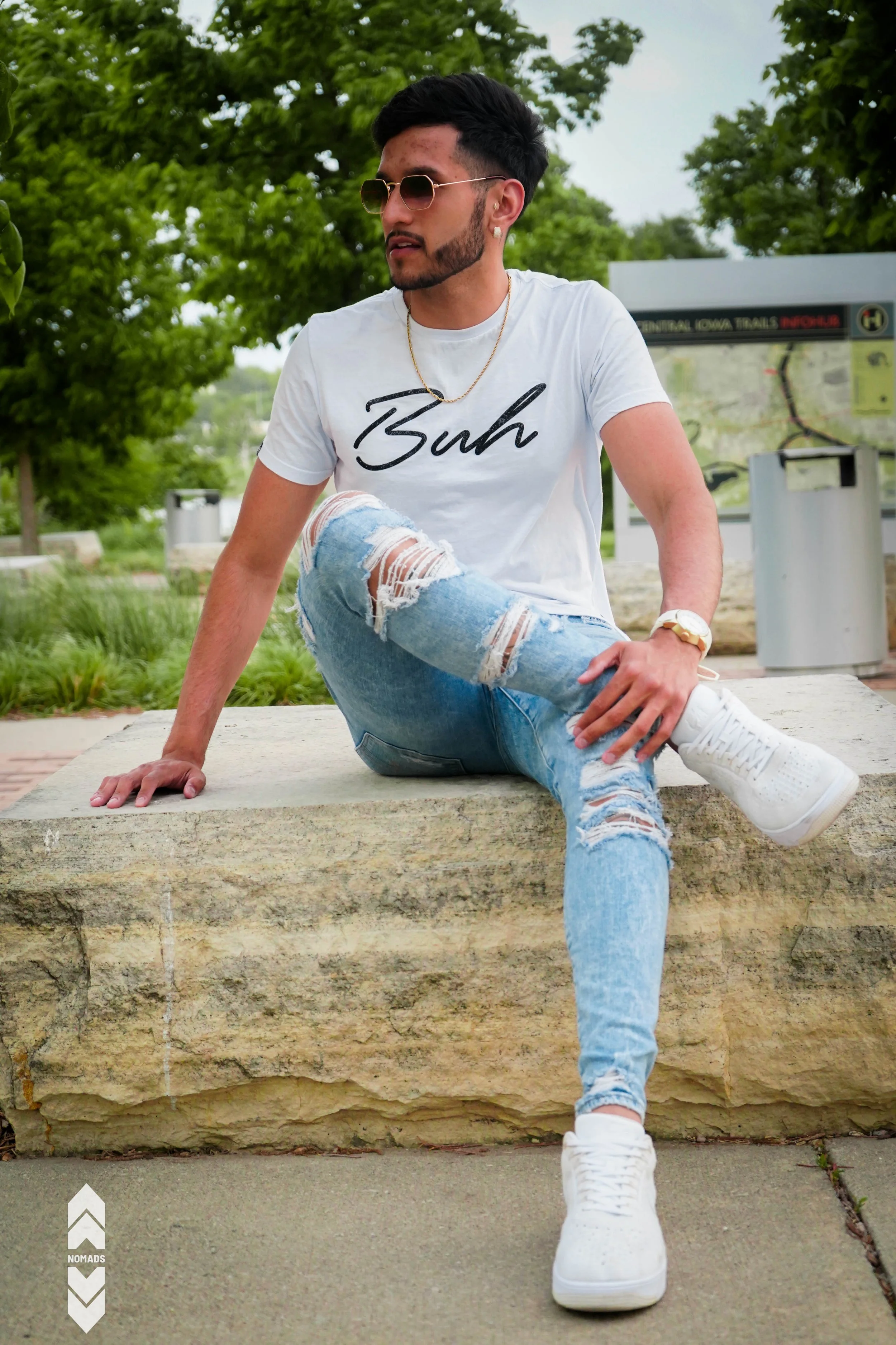 Young man sitting on a stone ledge outdoors, wearing sunglasses, a white t-shirt with black text, ripped jeans, and white sneakers, with green trees behind him.