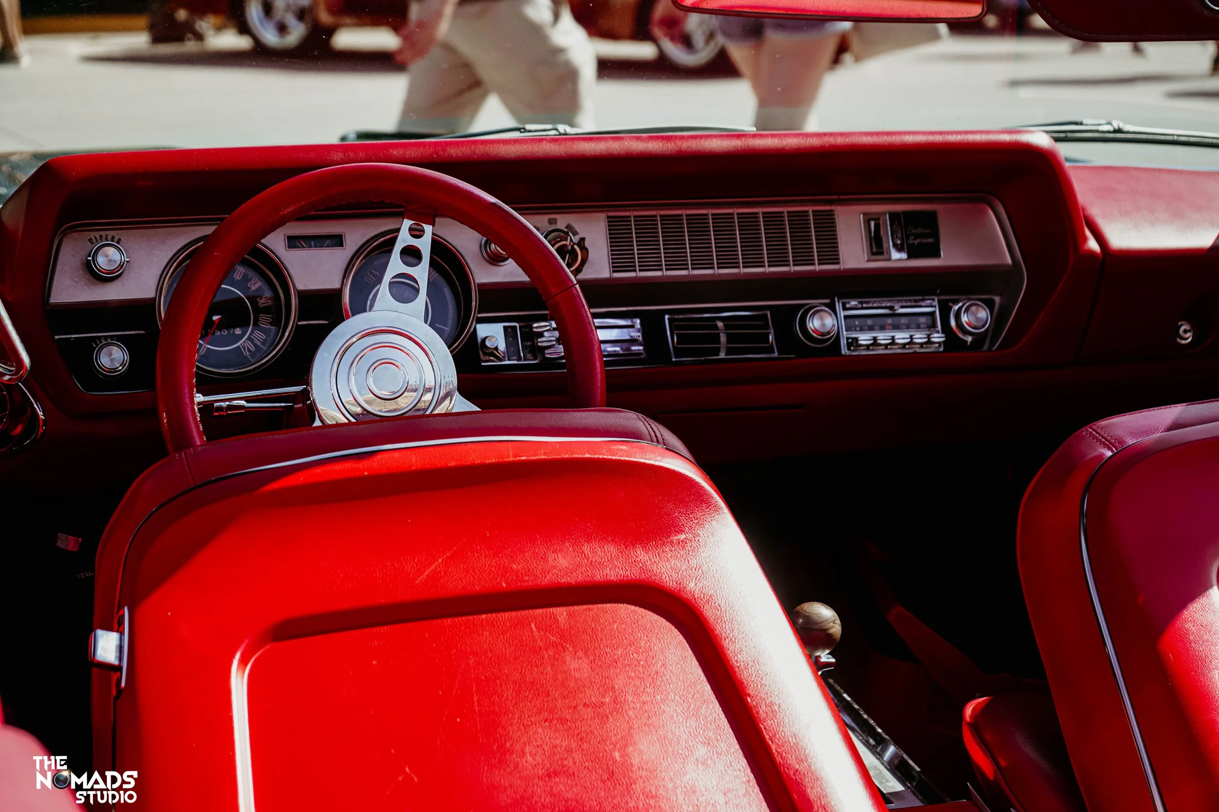 The image shows the interior dashboard of a vintage red car, with red upholstery and a steering wheel. The dashboard features analog gauges and control knobs, and the background includes a person walking by, seen through the windshield.