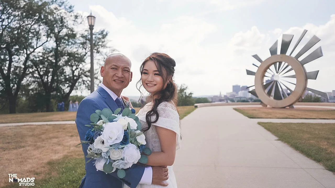 A bride and groom standing together outdoors on a paved path, holding a bouquet of white and blue flowers, with a large modern sculpture in the background.