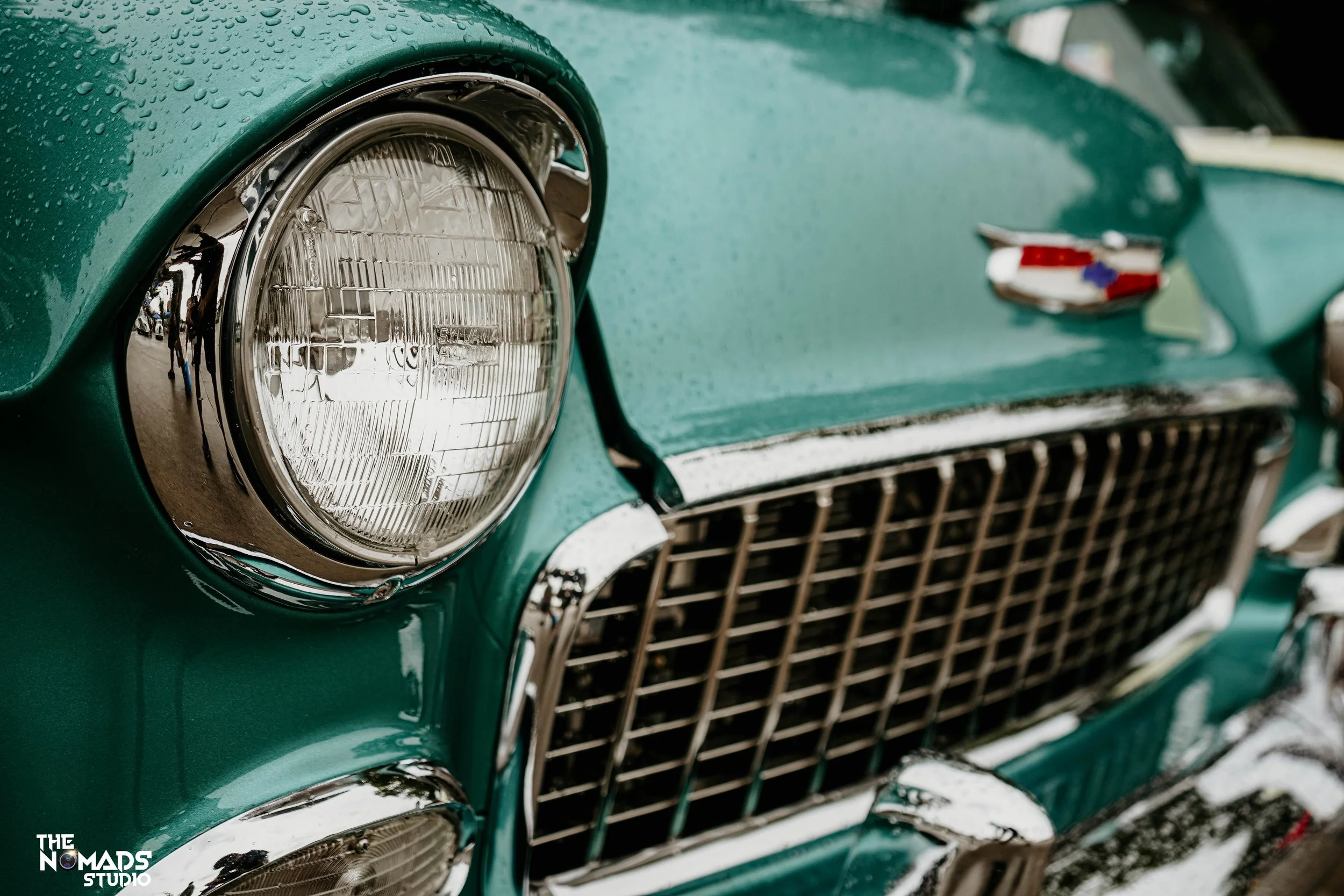 Close-up image of a vintage green car front, showing a headlight with a chrome rim, a grille below, and a badge on the hood, with water droplets on the surface.