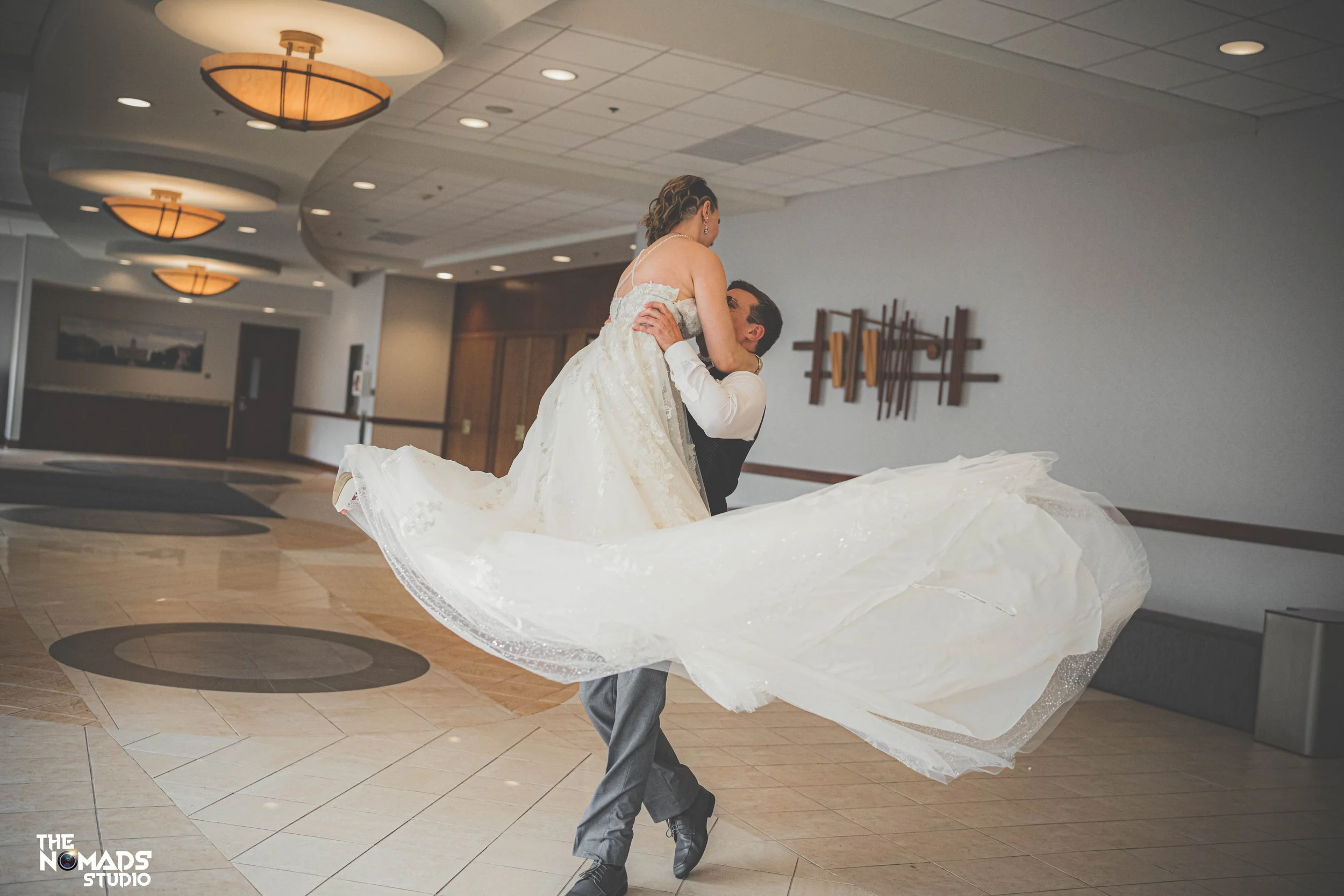 A man in gray formal pants and black shoes twirling a woman in a white wedding dress inside a modern room with a tiled floor and patterned wall art.