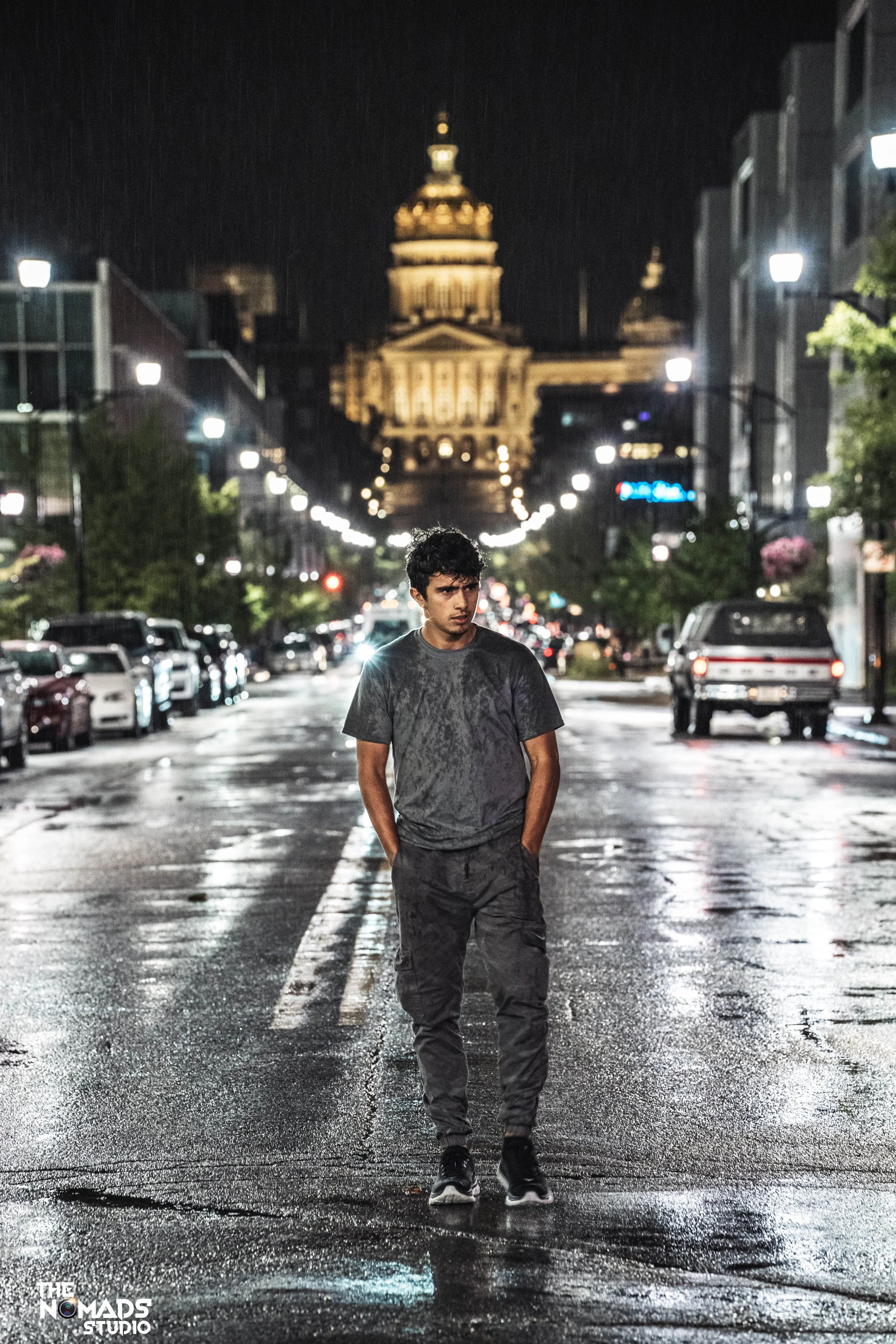 A young man in dark athletic clothing walks on a wet city street at night, with a large illuminated building in the background and streetlights lining the street.
