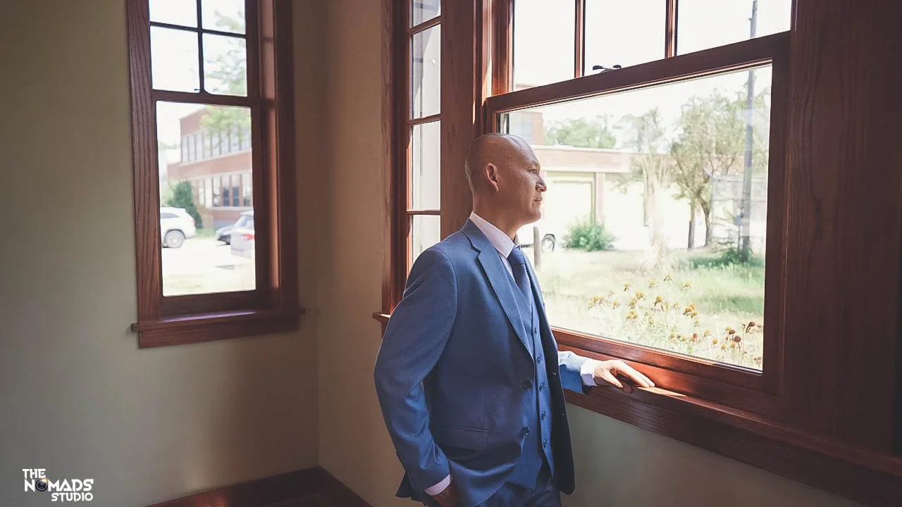 A man in a blue suit looking out of a large window with a view of trees and a building outside.