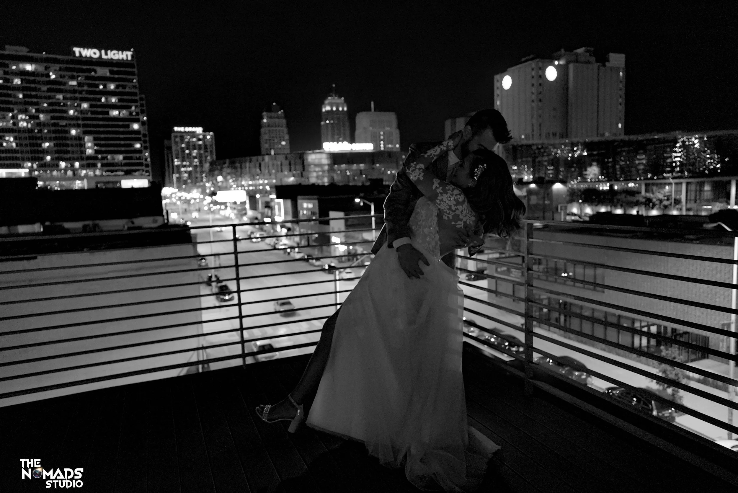 A black and white photo of a couple, with the man lifting the woman, on a balcony at night. City skyline with tall buildings and bright lights in the background.