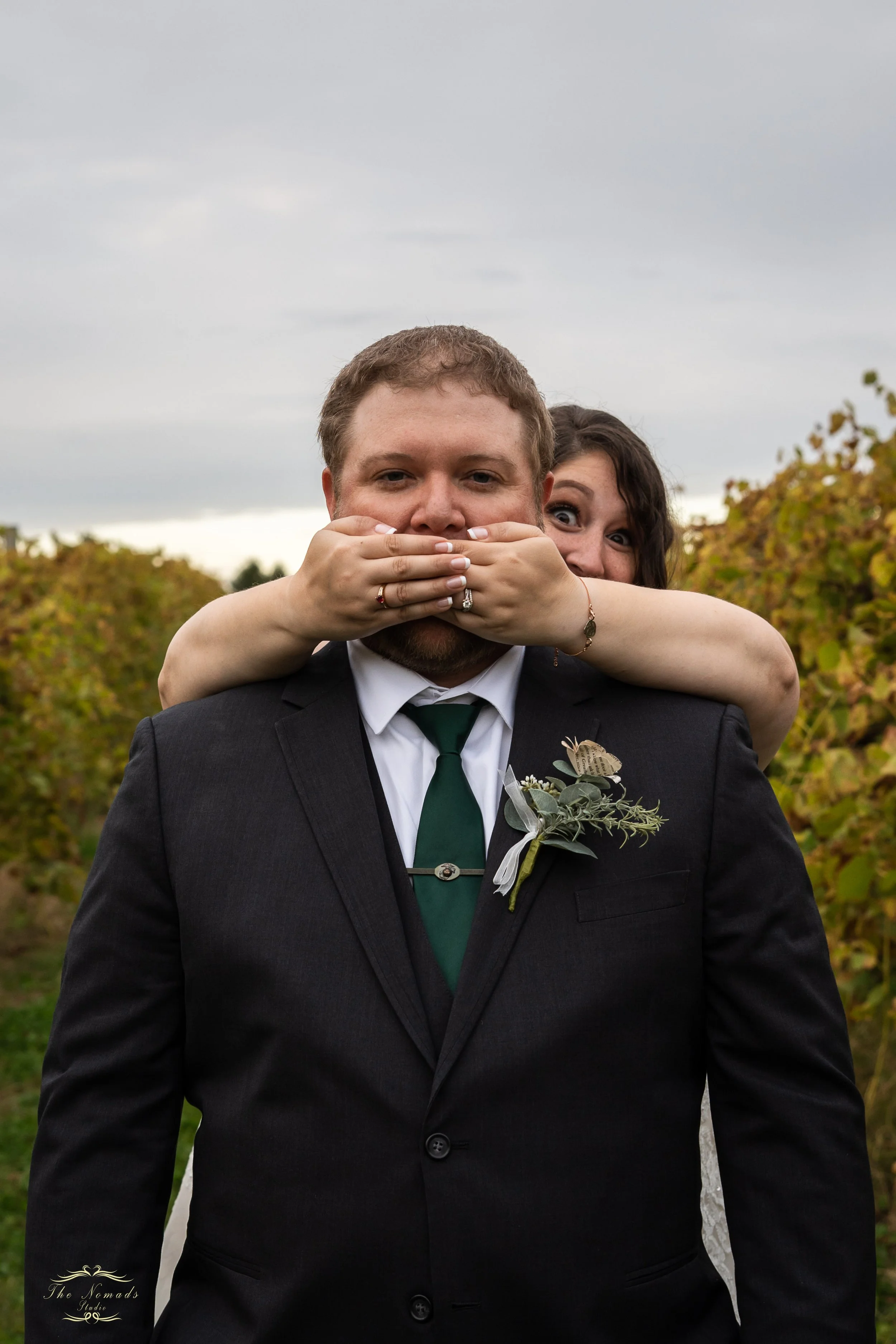 A couple in formal attire standing outdoors with the woman covering the man's eyes with her hands. The man is wearing a dark suit with a white shirt, green tie, and boutonniere. The woman is behind him, with brown hair and wide eyes, smiling. The setting appears to be a vineyard with rows of grapevines and an overcast sky.