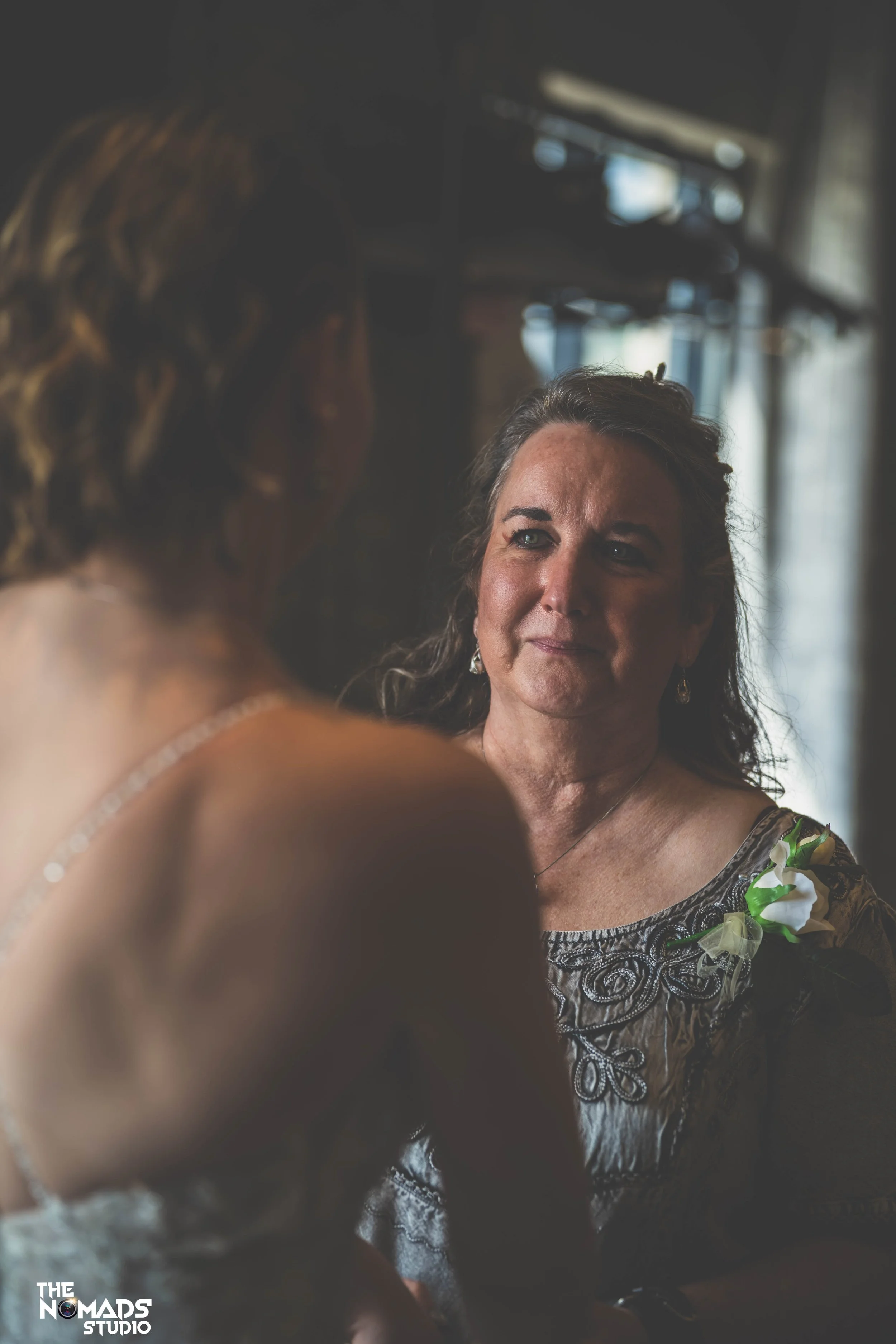 A woman with curly hair smiling and looking at another person whose back is to the camera. The woman is dressed in a patterned blouse and has a corsage on her shoulder. The background is blurred with natural light entering through windows.
