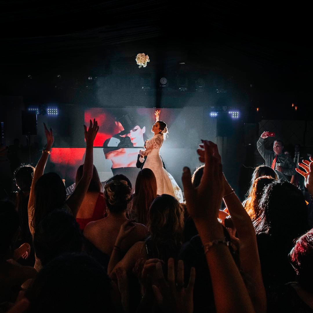 A wedding reception with a bride in a wedding dress performing a dance on stage in front of an audience, with hands raised in celebration and a large screen behind her displaying an image.