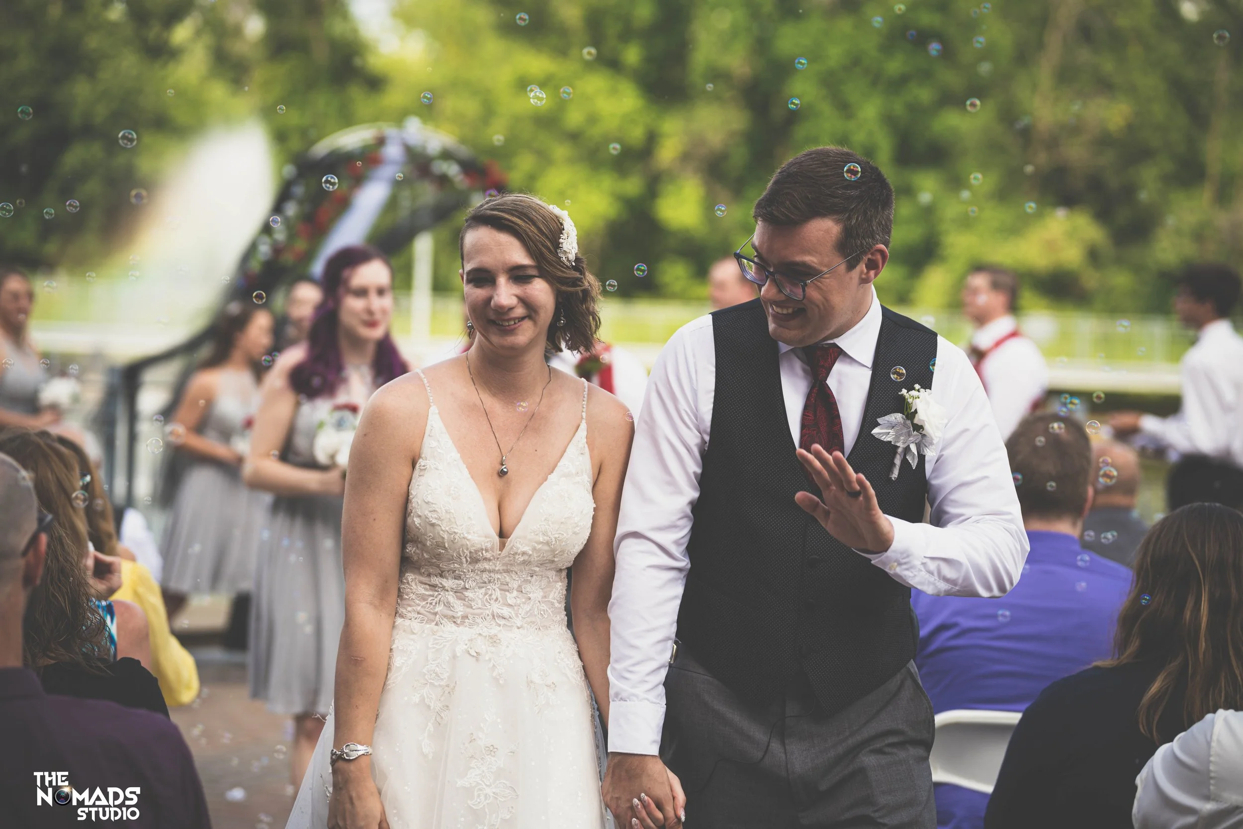 A bride and groom holding hands during their wedding ceremony outdoors, surrounded by friends and family with trees and bubbles in the background.