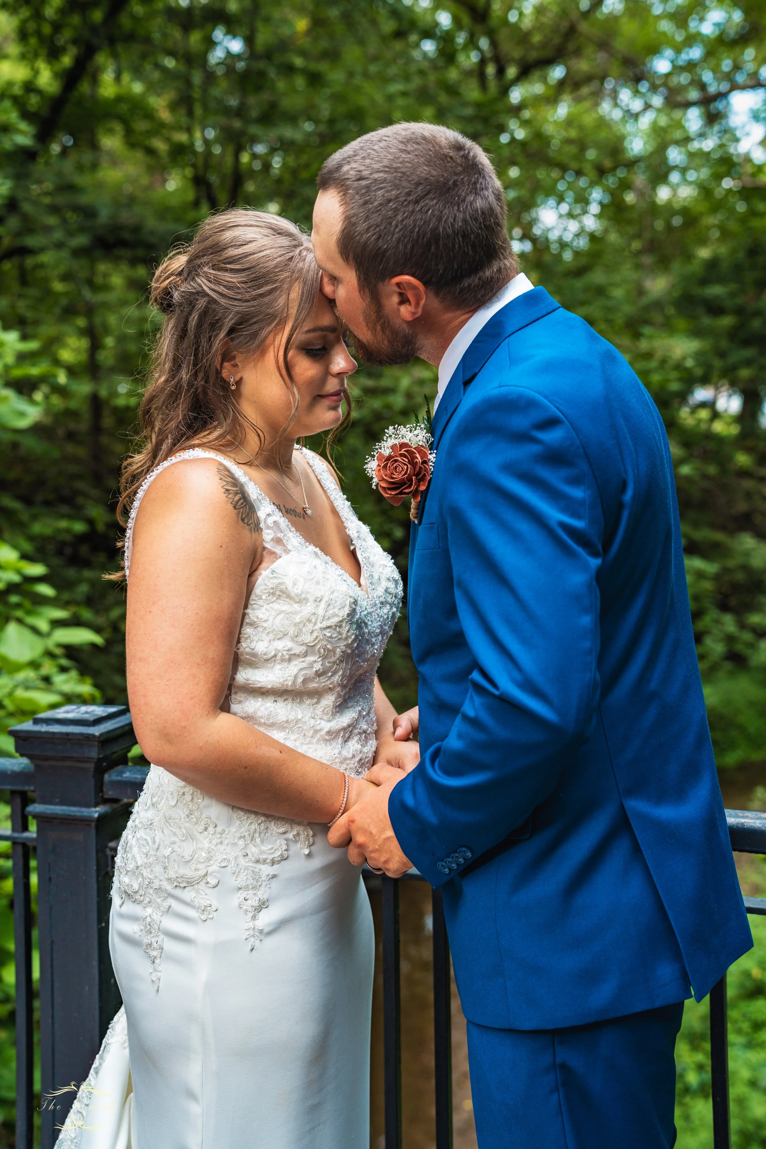 A couple in wedding attire holding hands and sharing a kiss outdoors in a wooded area.