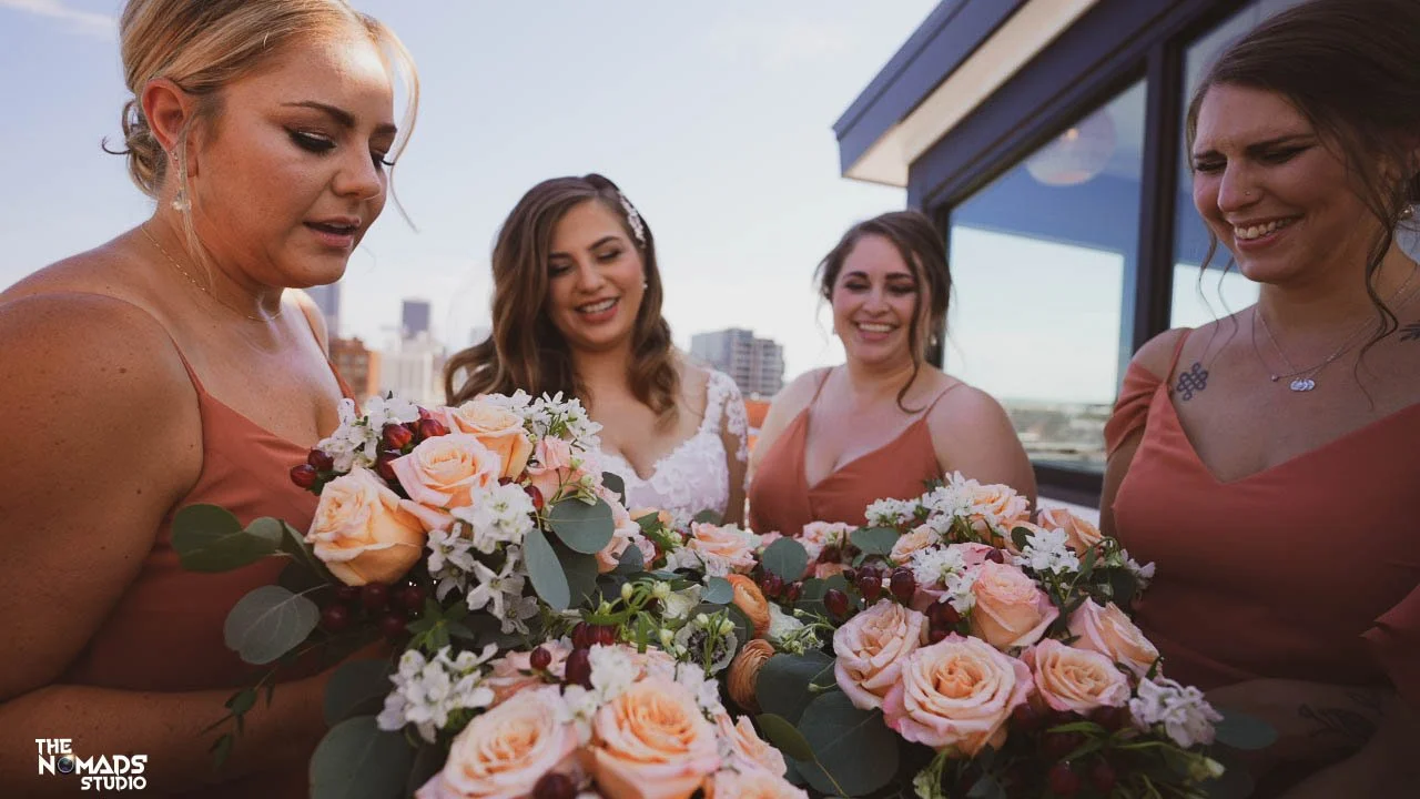 Four women dressed in formal attire holding a large wedding bouquet of pink roses and white flowers, standing outside near a building with cityscape in the background.