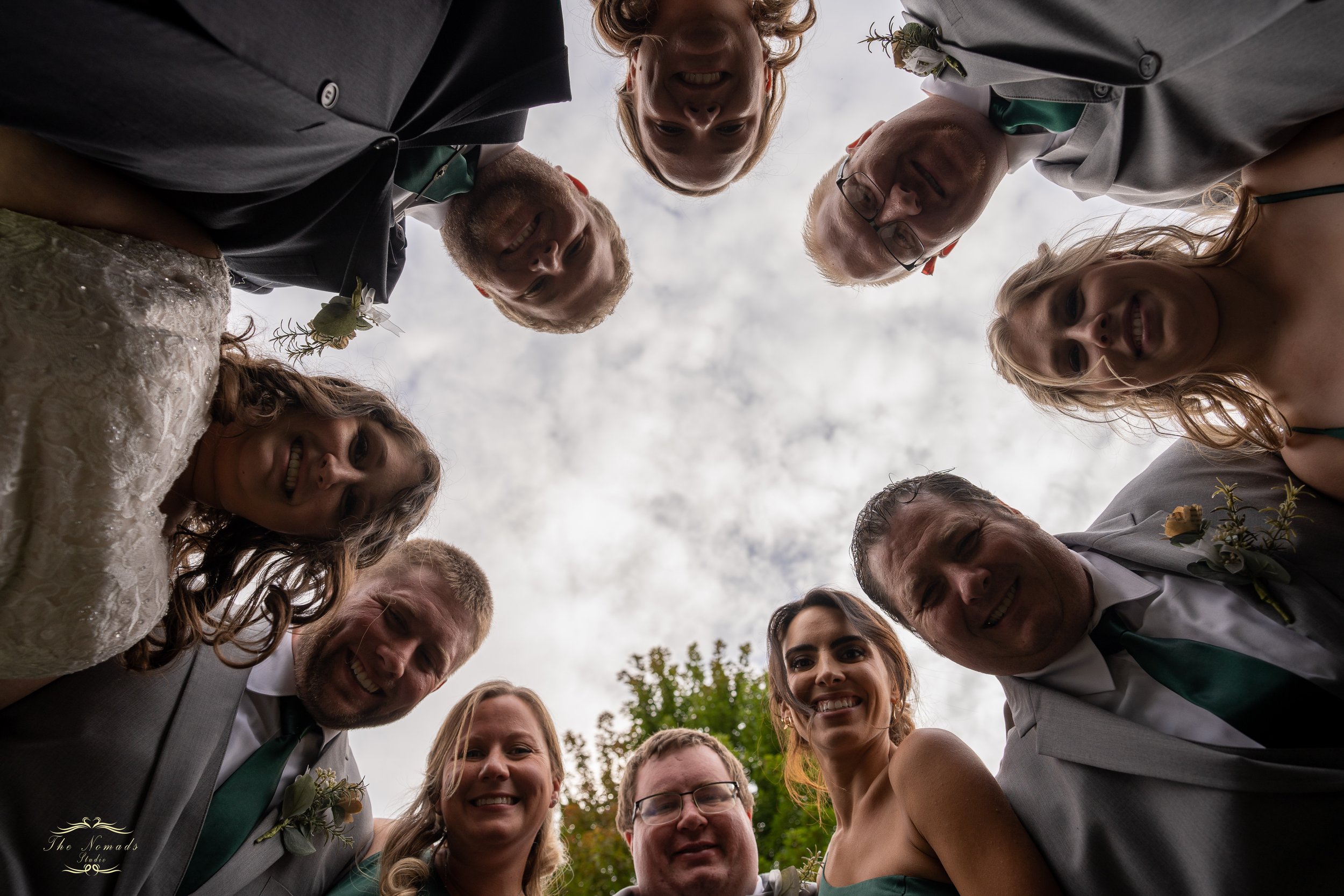 Group of smiling people gathered in a circle looking down at the camera, taken from below, outdoors with cloudy sky and trees in the background.