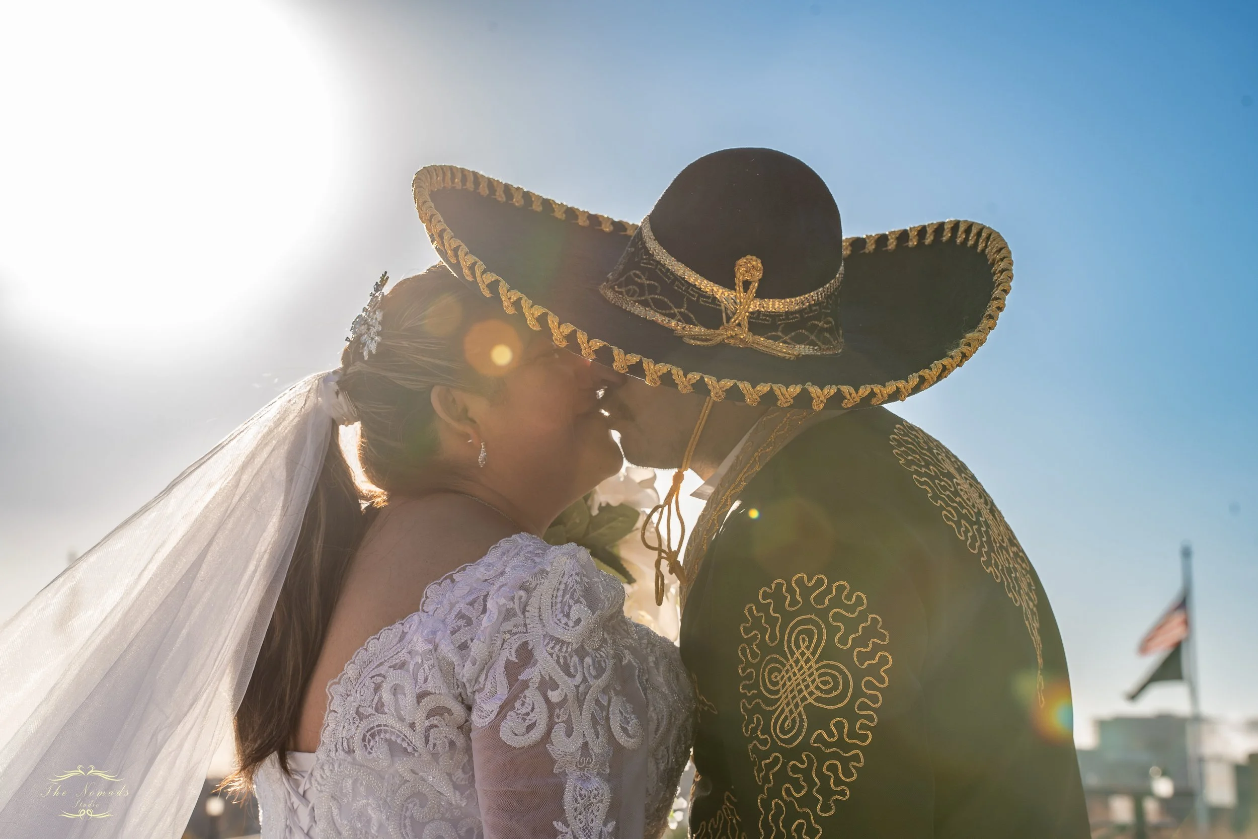 A bride and groom sharing a kiss outdoors, with the bride wearing a white lace wedding dress and veil, and the groom wearing a black traditional Mexican sombrero with gold embroidery, under a bright sun and blue sky.
