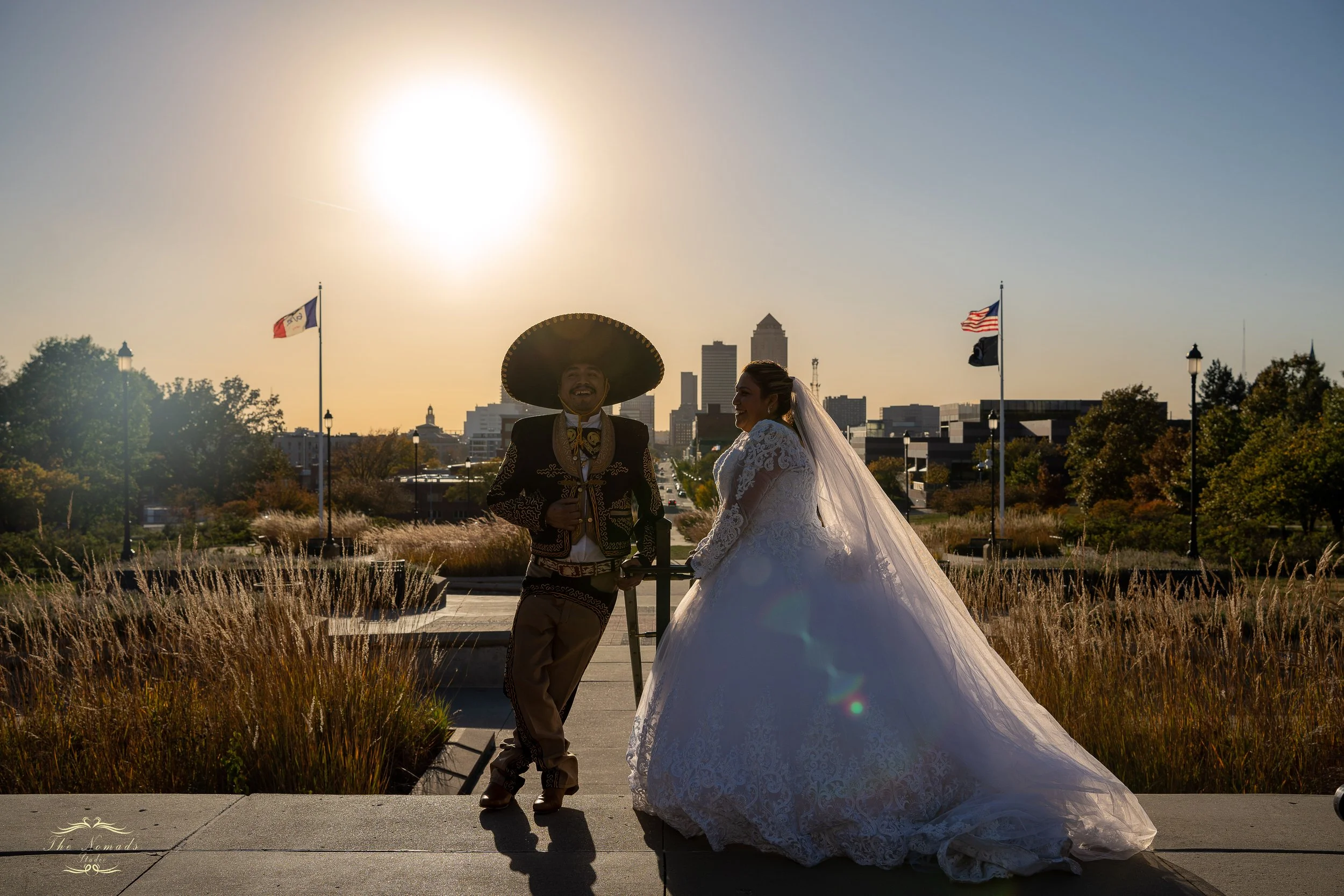 A bride and groom stand together outdoors at sunset, dressed in wedding attire, with a city skyline in the background and flags on poles nearby.