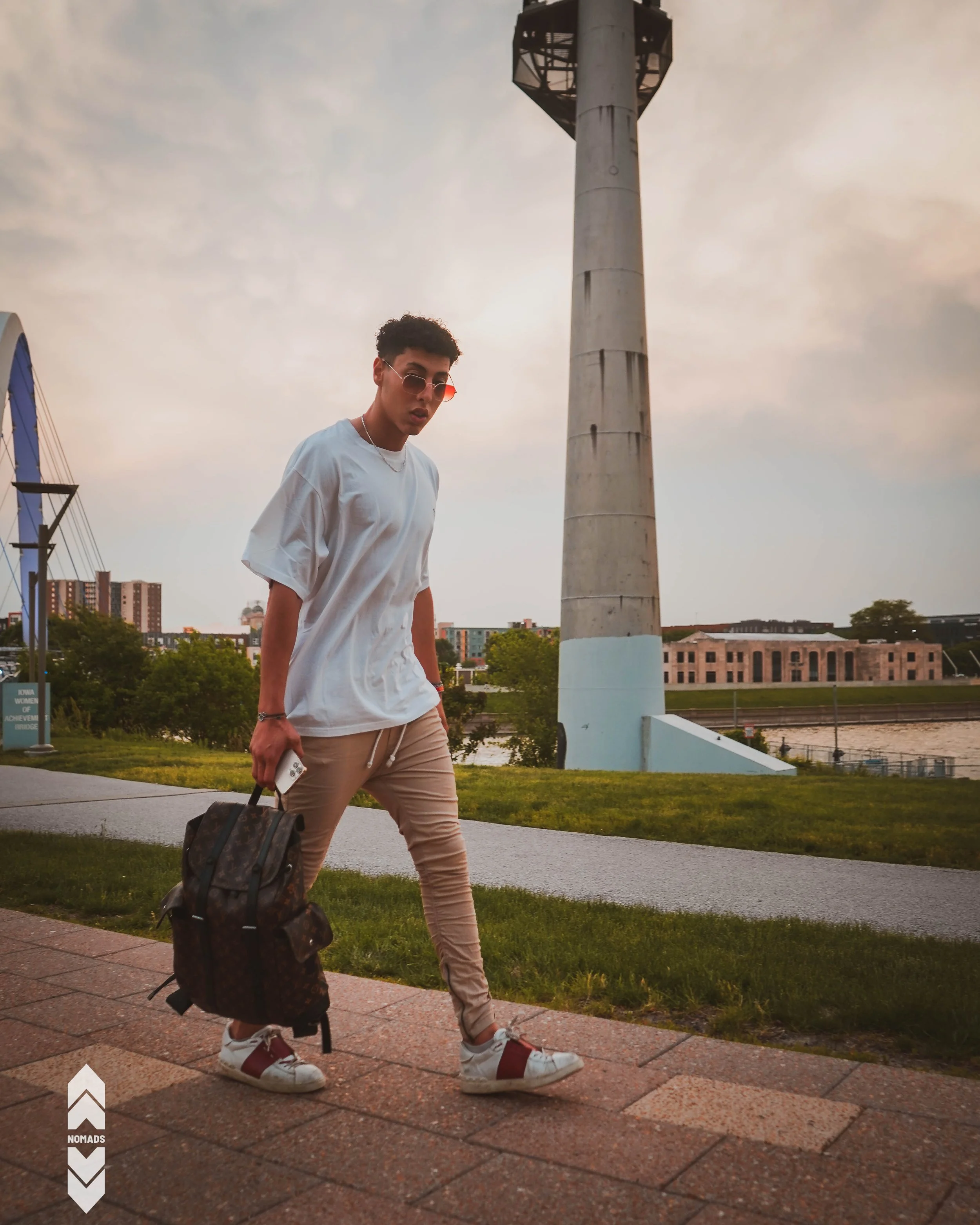 A young man wearing sunglasses, a white T-shirt, beige pants, and white sneakers with red straps, holding a phone and a backpack, walking on a sidewalk in an urban park during dusk.