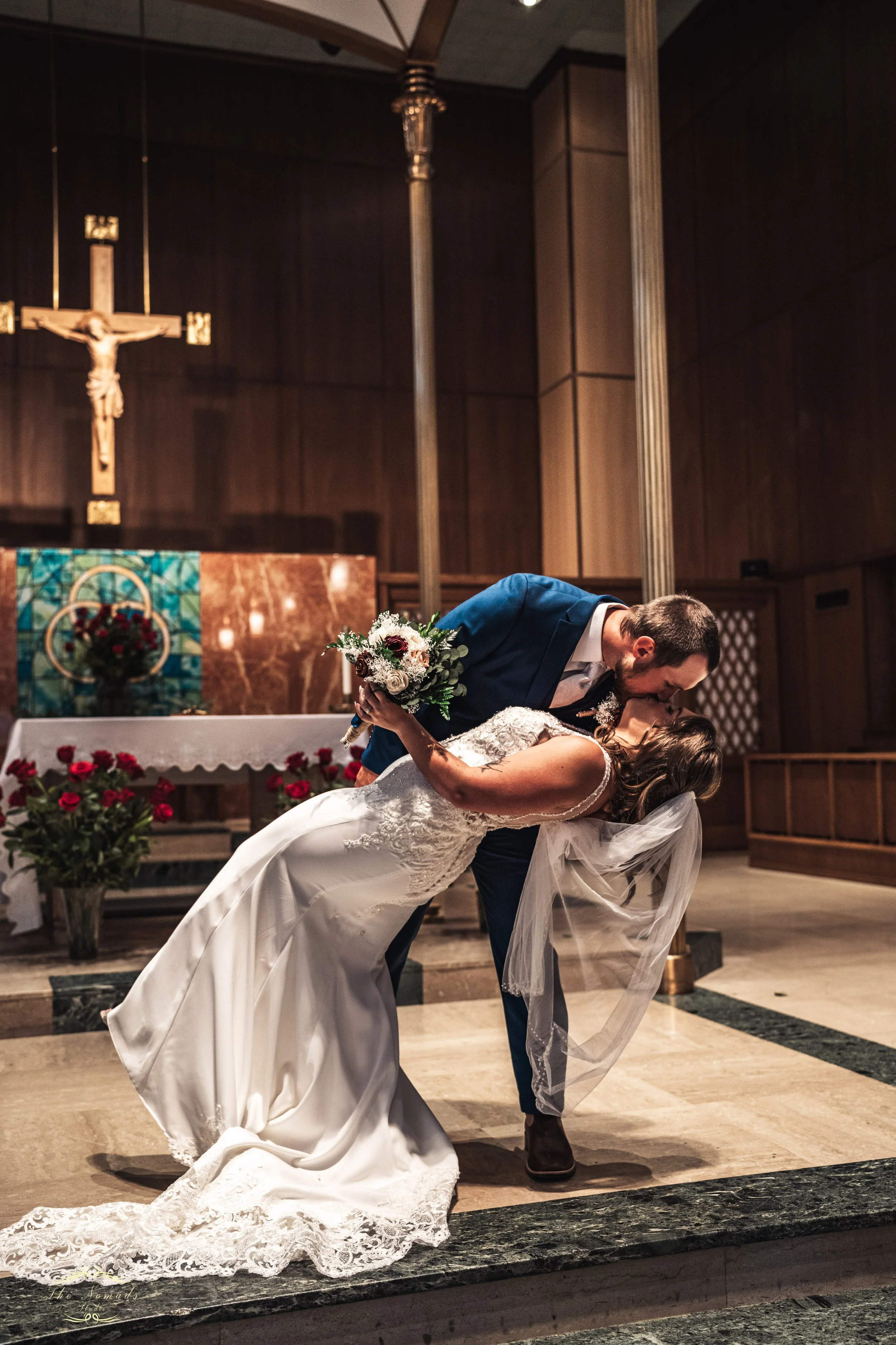 A man in a blue suit and brown boots is kissing a woman in a wedding dress who is bending backwards in a church setting. The woman holds a bouquet of flowers, and the church altar in the background features crucifix and religious decor, with a dark w