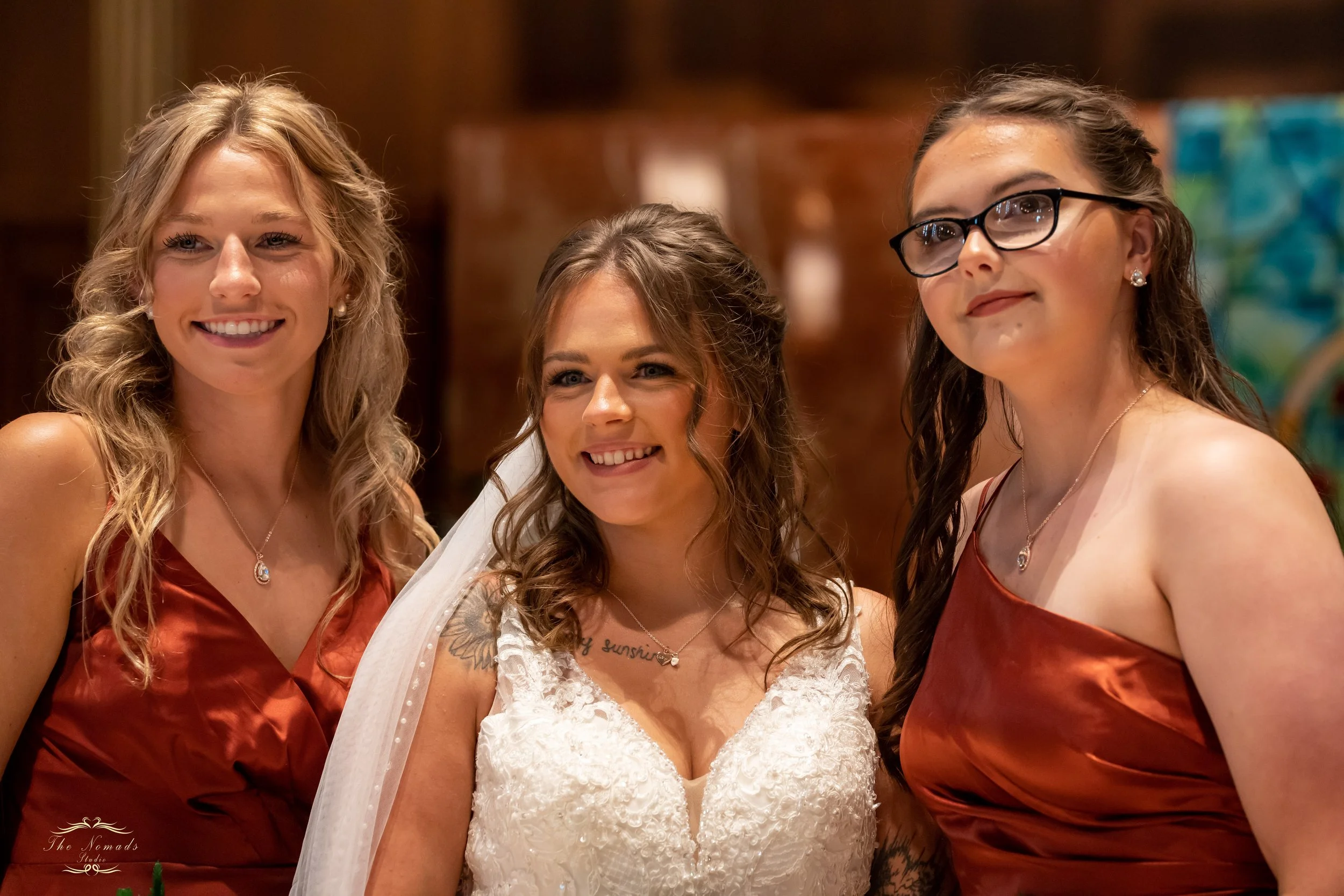 Three women at a wedding celebration, with the bride in the center wearing a white lace dress and veil, and two bridesmaids on bust, wearing matching reddish satin dresses, all smiling.