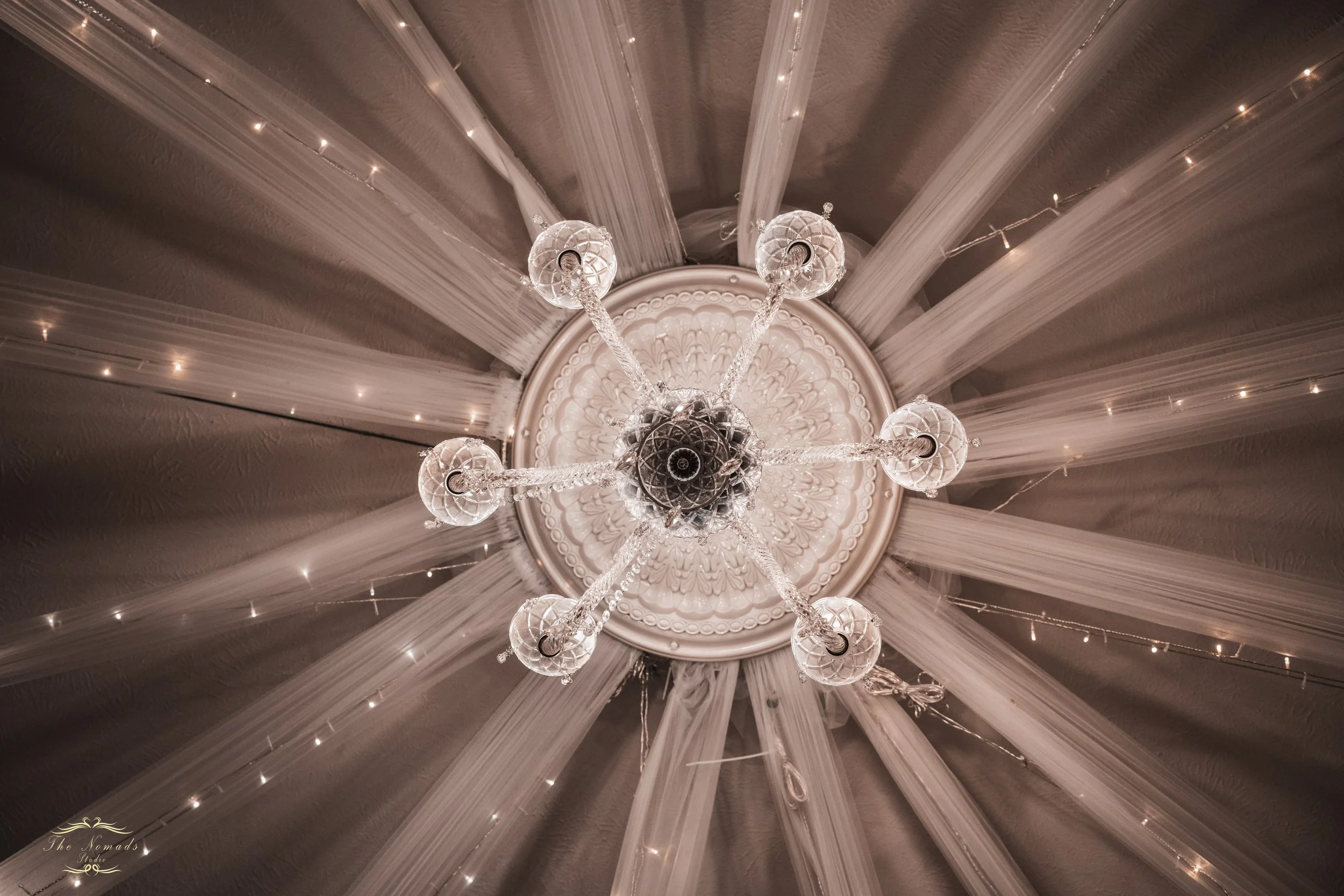 A close-up of a chandelier with multiple glass globes hanging from a decorative ceiling medallion, with the ceiling decorated with strings of small lights and flowing fabric or ribbons.
