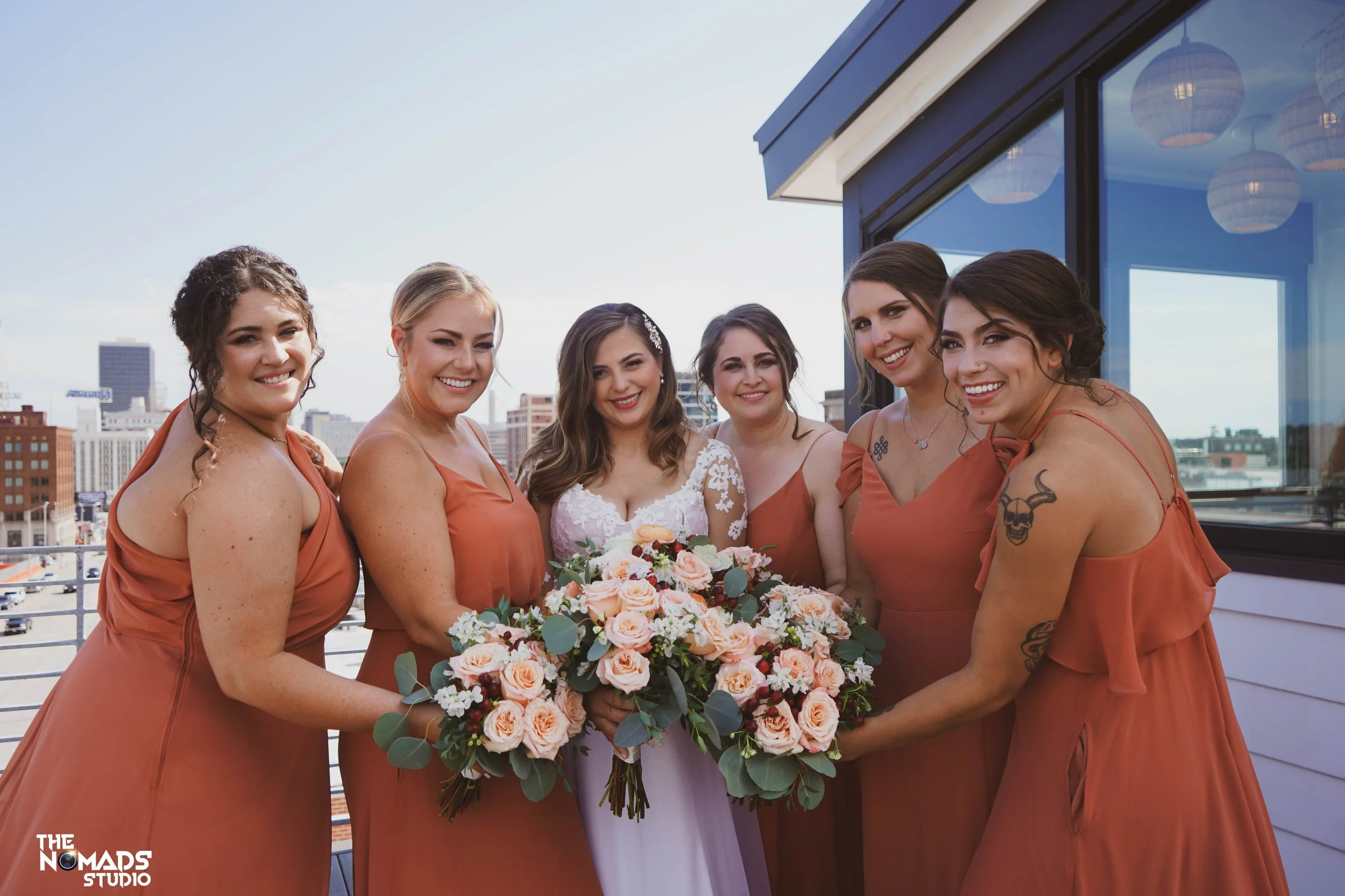 A group of seven women dressed in matching rust-colored dresses, standing on a rooftop with a city skyline in the background. One woman in the center is holding a bouquet of pink, white, and cream flowers. They are smiling and posing for a photo.