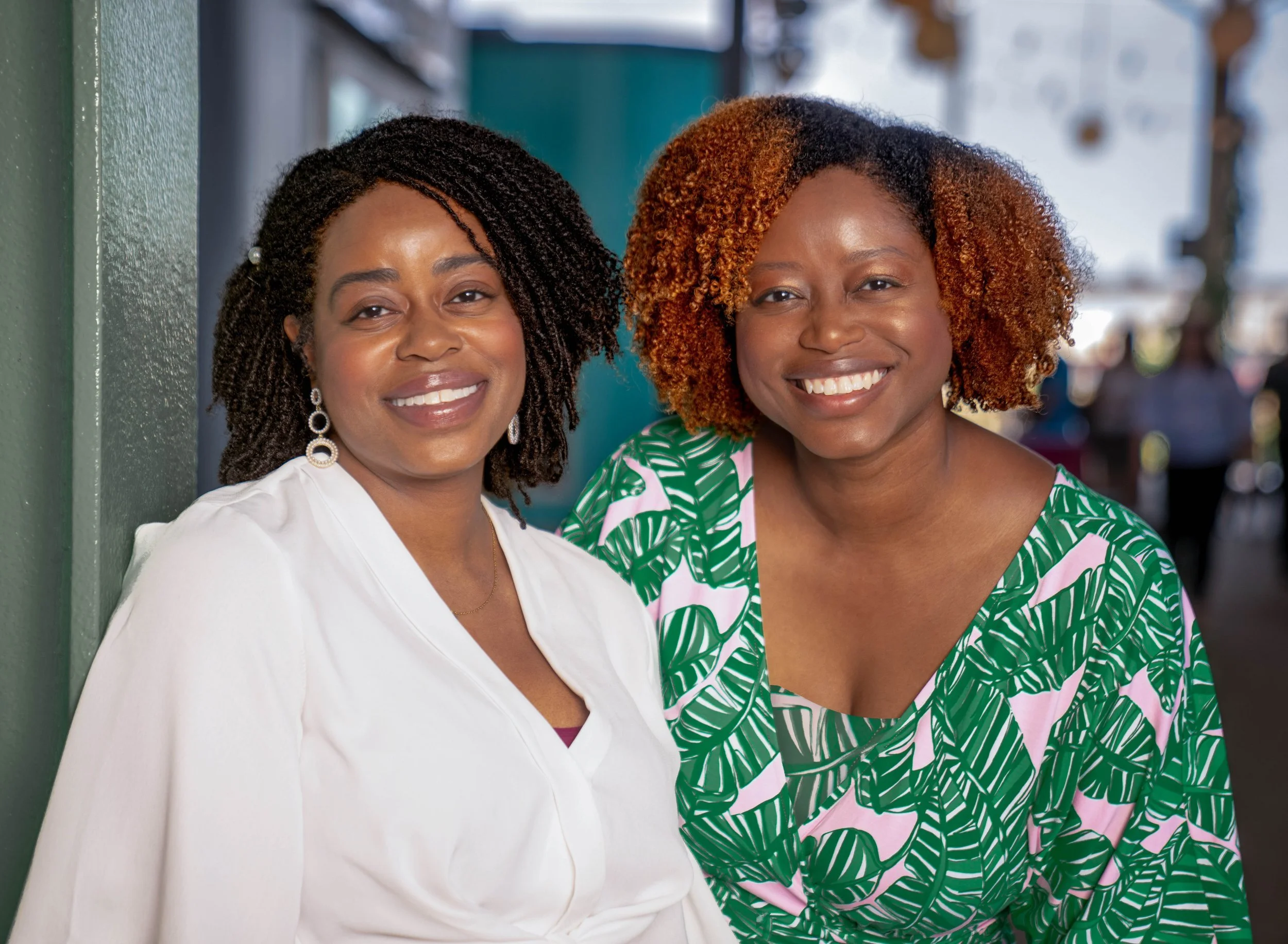 Two women standing close together and smiling at the camera, one wearing a white blouse with earrings and the other in a green and pink patterned dress with curly hair, in an outdoor setting with people in the background.