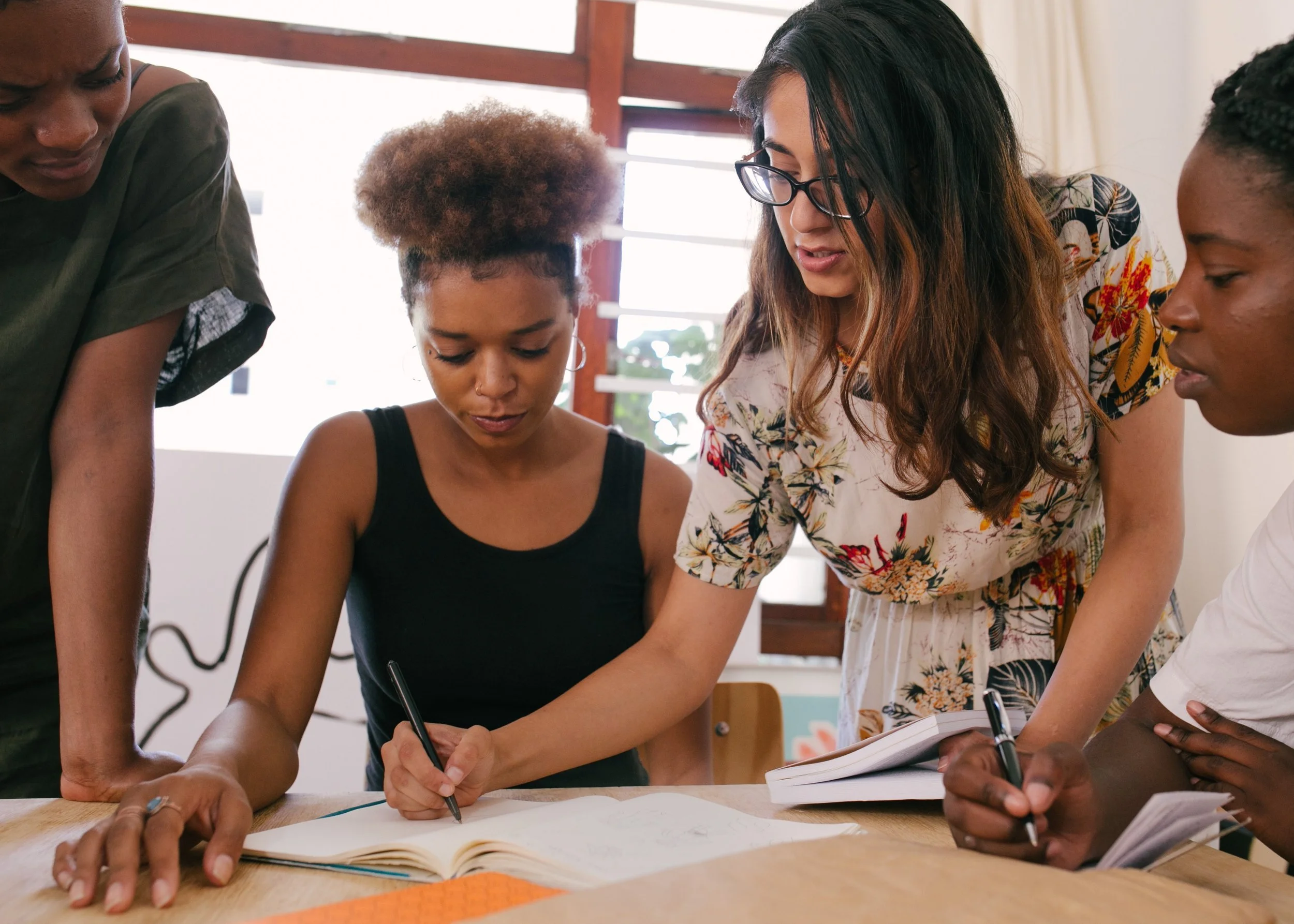 A diverse group of women engaged in a collaborative discussion at a table, with some writing in notebooks.