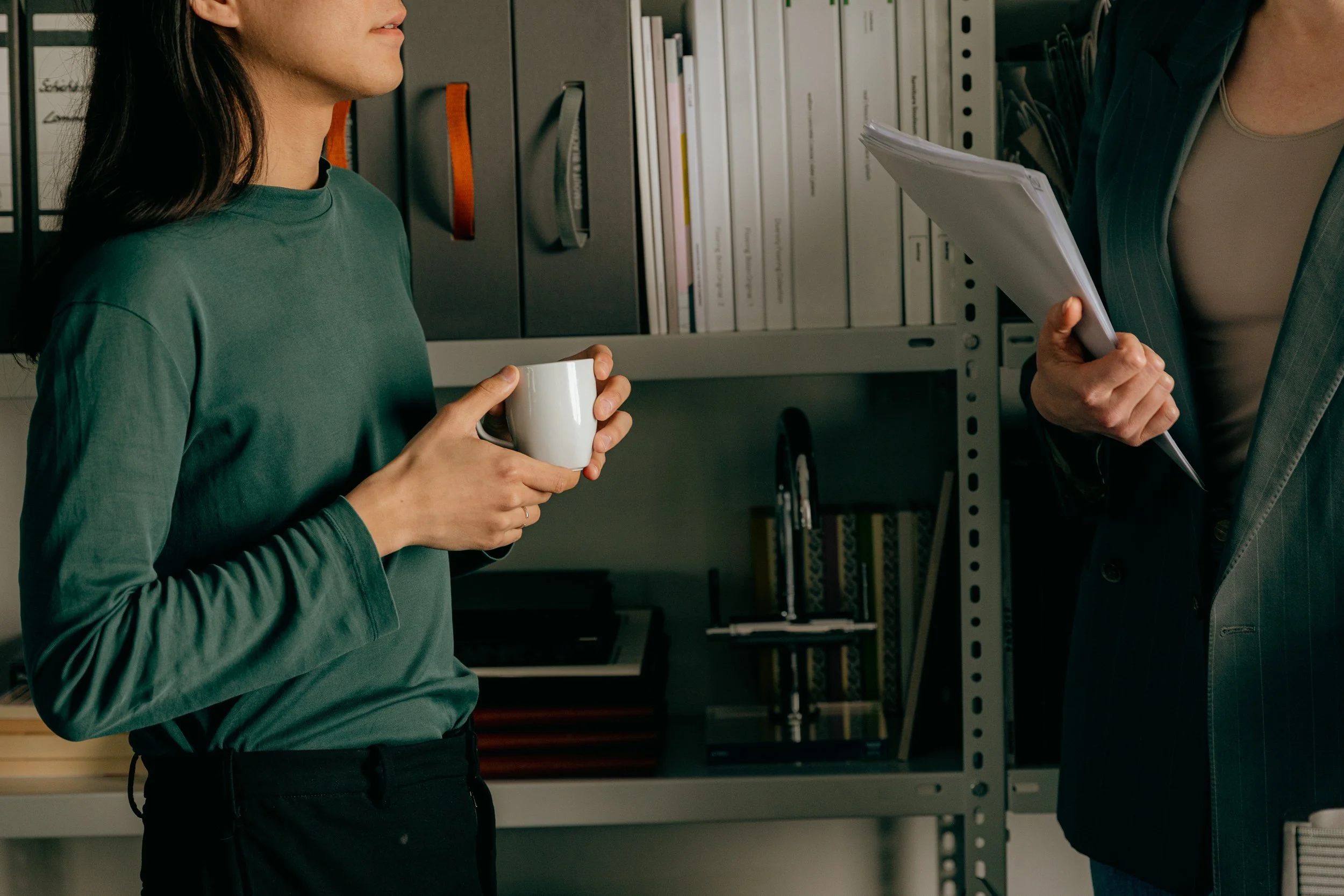 Two people in a conversation in an office, one holding a white mug, and the other holding a folder of papers.