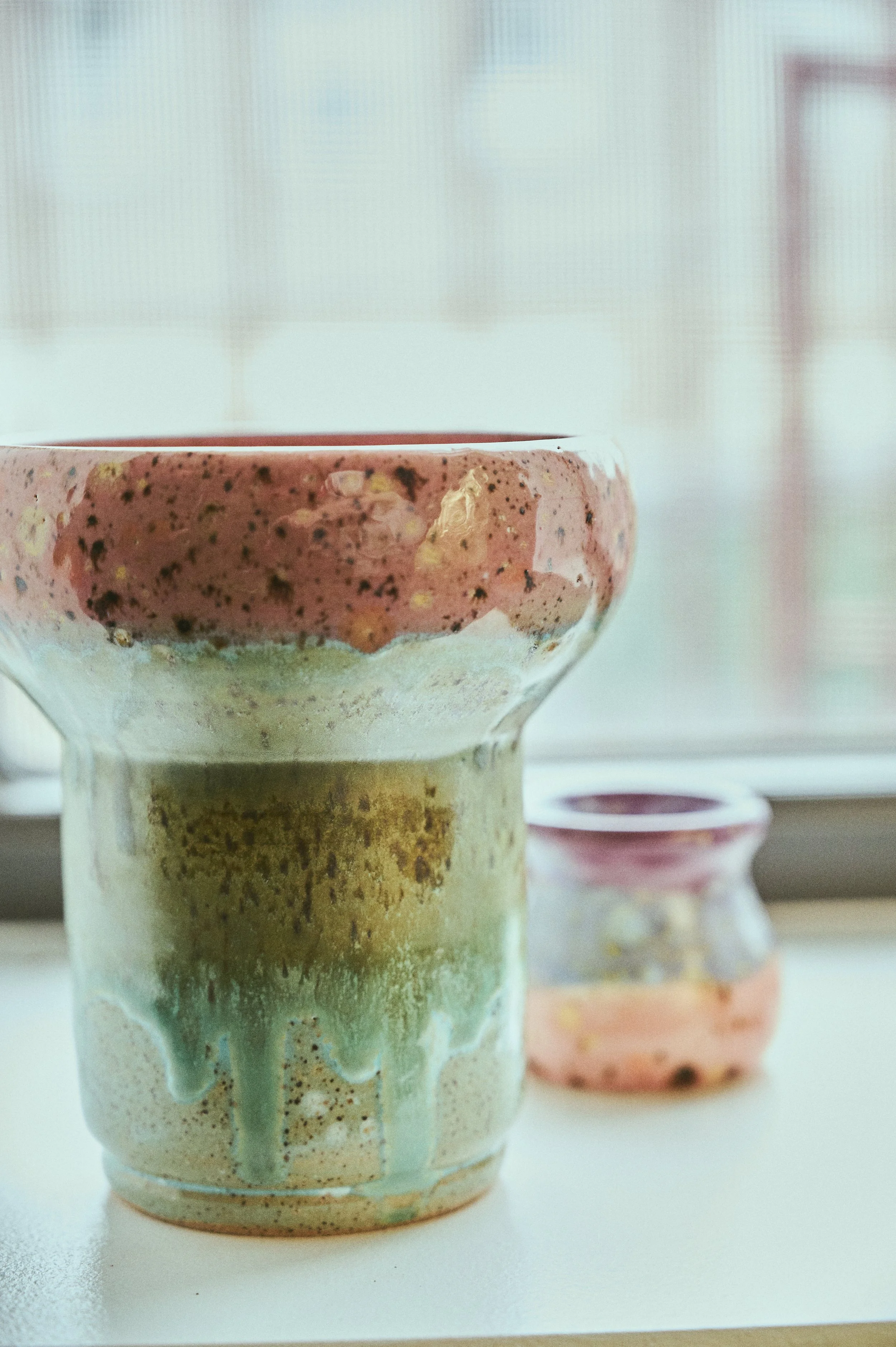 Close-up of a ceramic glaze with a speckled glaze, showing drips of glaze running down the side. In the background, additional ceramic mini vases with similar glazing