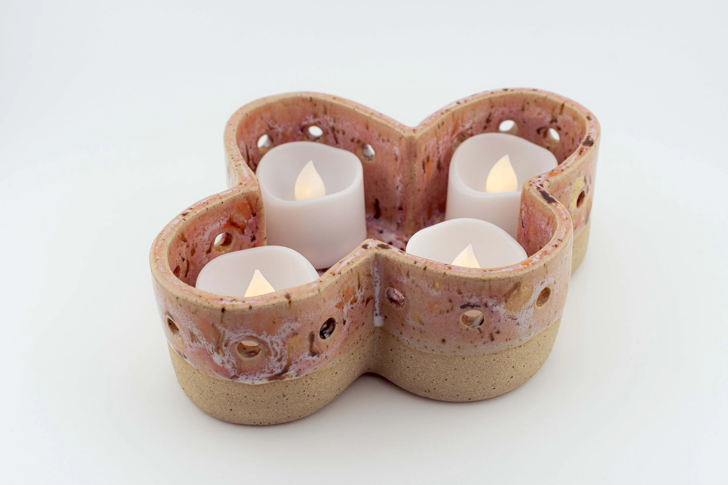 A heart-shaped candle holder made of pink and beige ceramic with small holes, holding three white candle flameless candles burning.