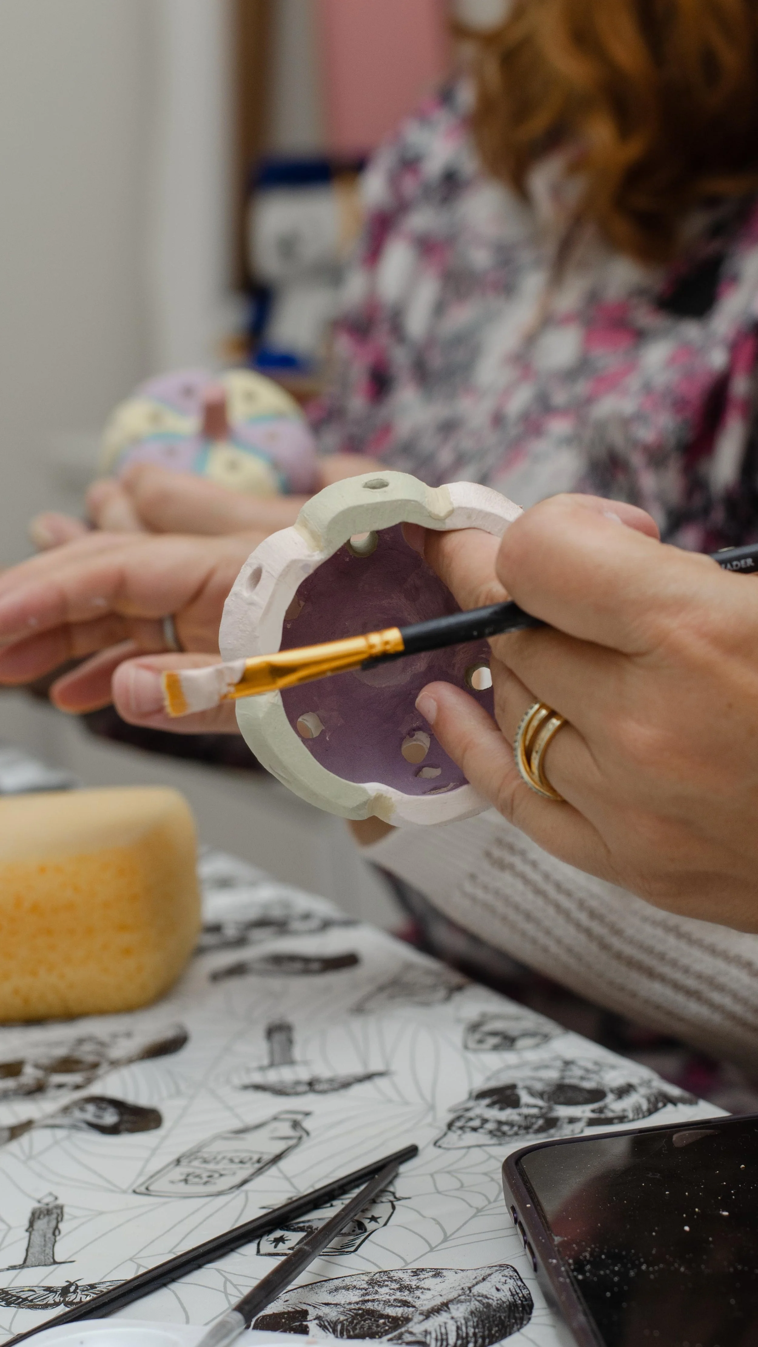 A person painting a colorful ceramic or clay object with a paintbrush at a table. Other art supplies and sketches are visible on the table.