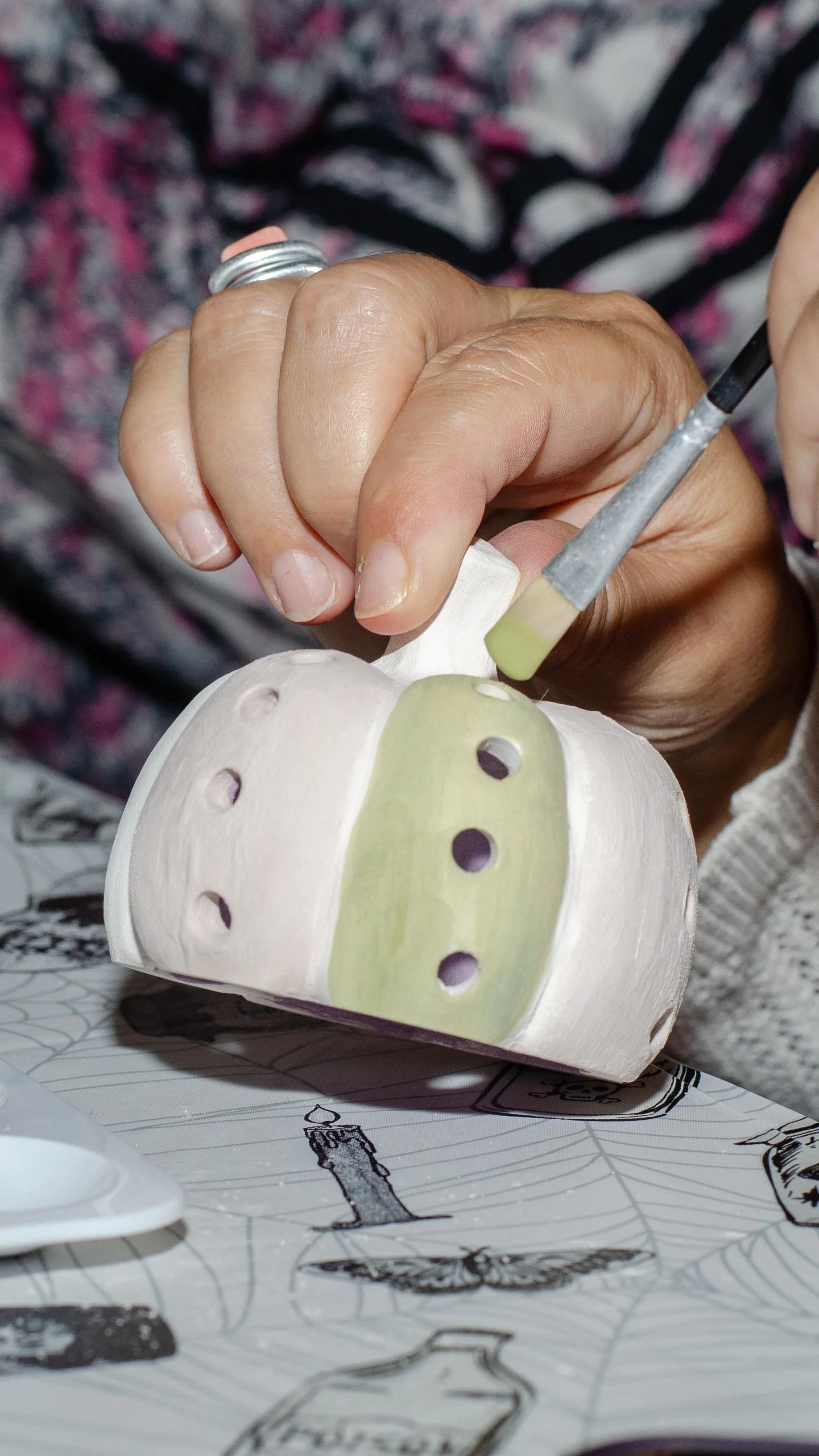 A person painting a ceramic object, using a small brush, with holes and painted green details. The background shows a patterned cloth, and the surface has black and white sketches or illustrations.