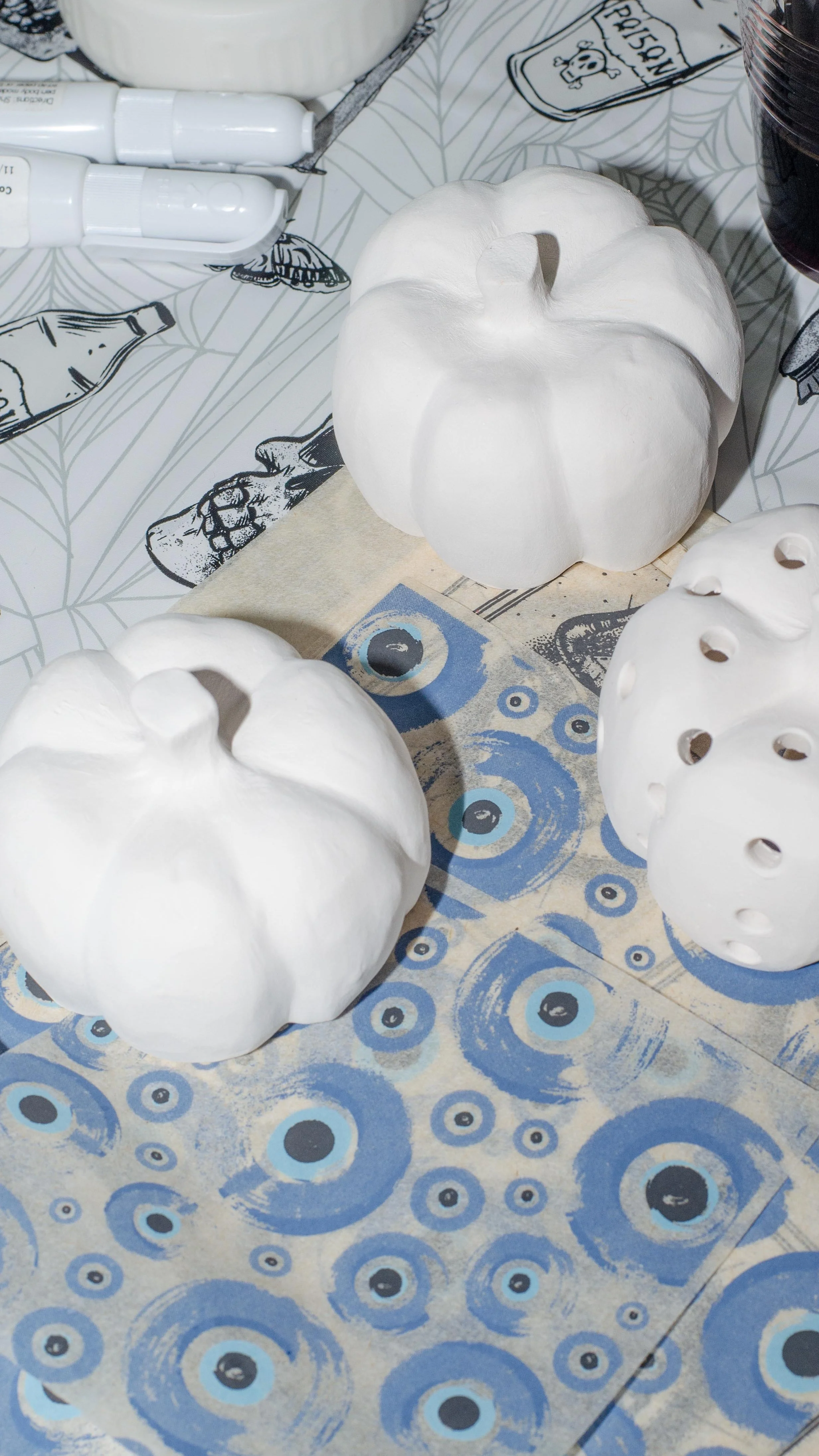 White ceramic pumpkins on a Halloween-themed tablecloth, with bottles of white paint or glue nearby.
