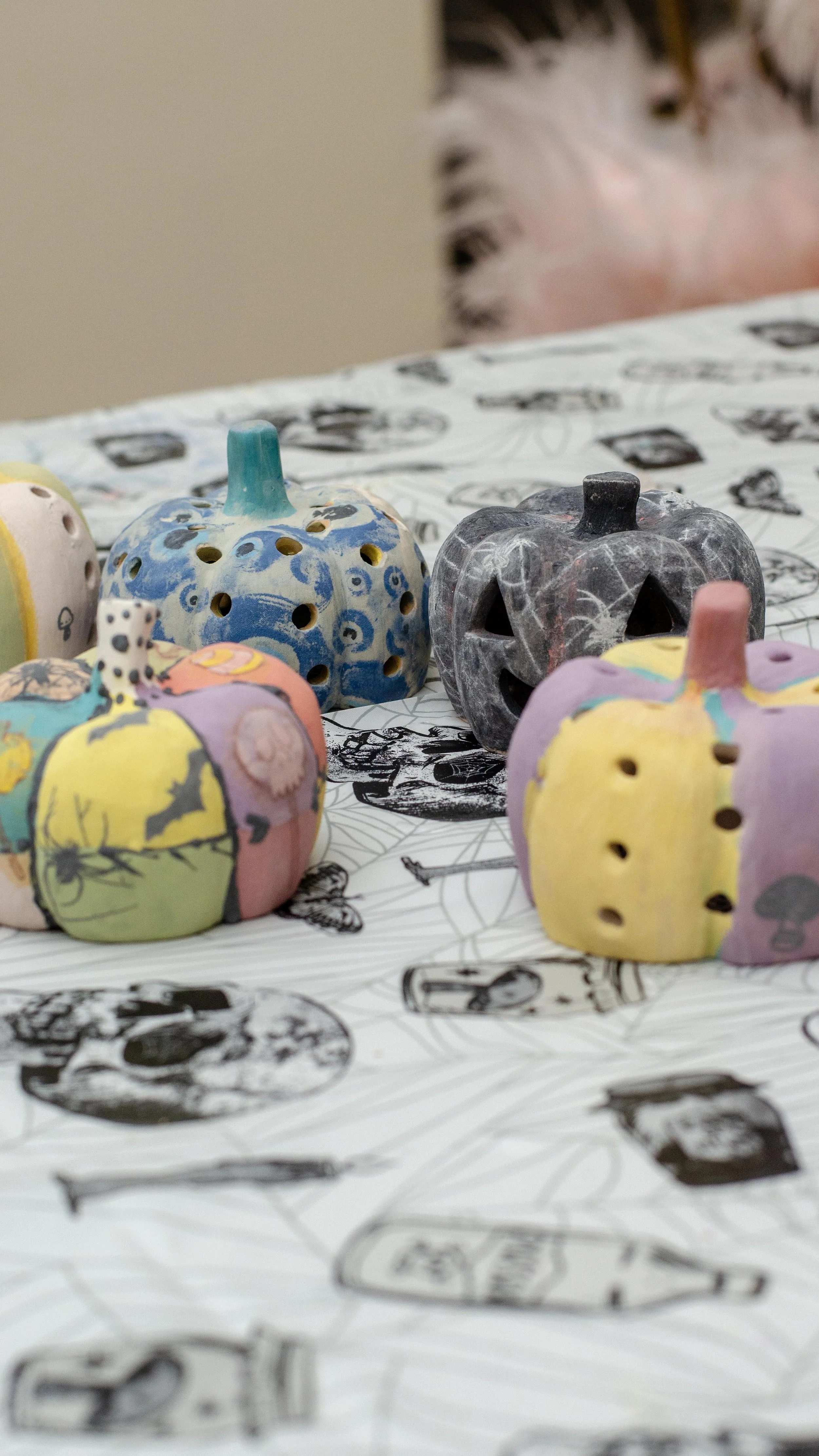 Colorful ceramic pumpkins and a black carved pumpkin on a printed tablecloth with black and white drawings.