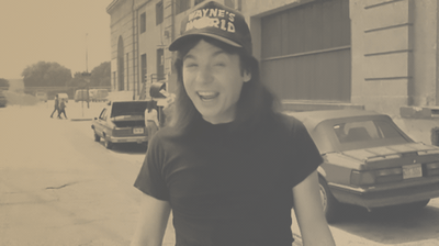 A man smiling and wearing a cap that says 'Wayne's World' standing on a city street with parked cars and buildings.