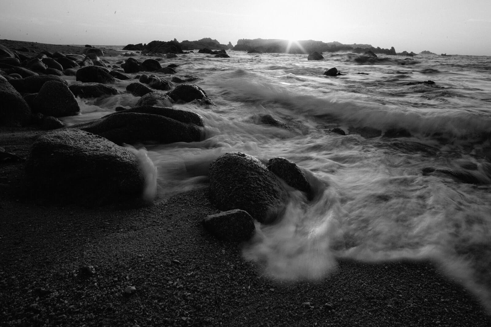 Black and white photo of the ocean rushing over rocks at sunset