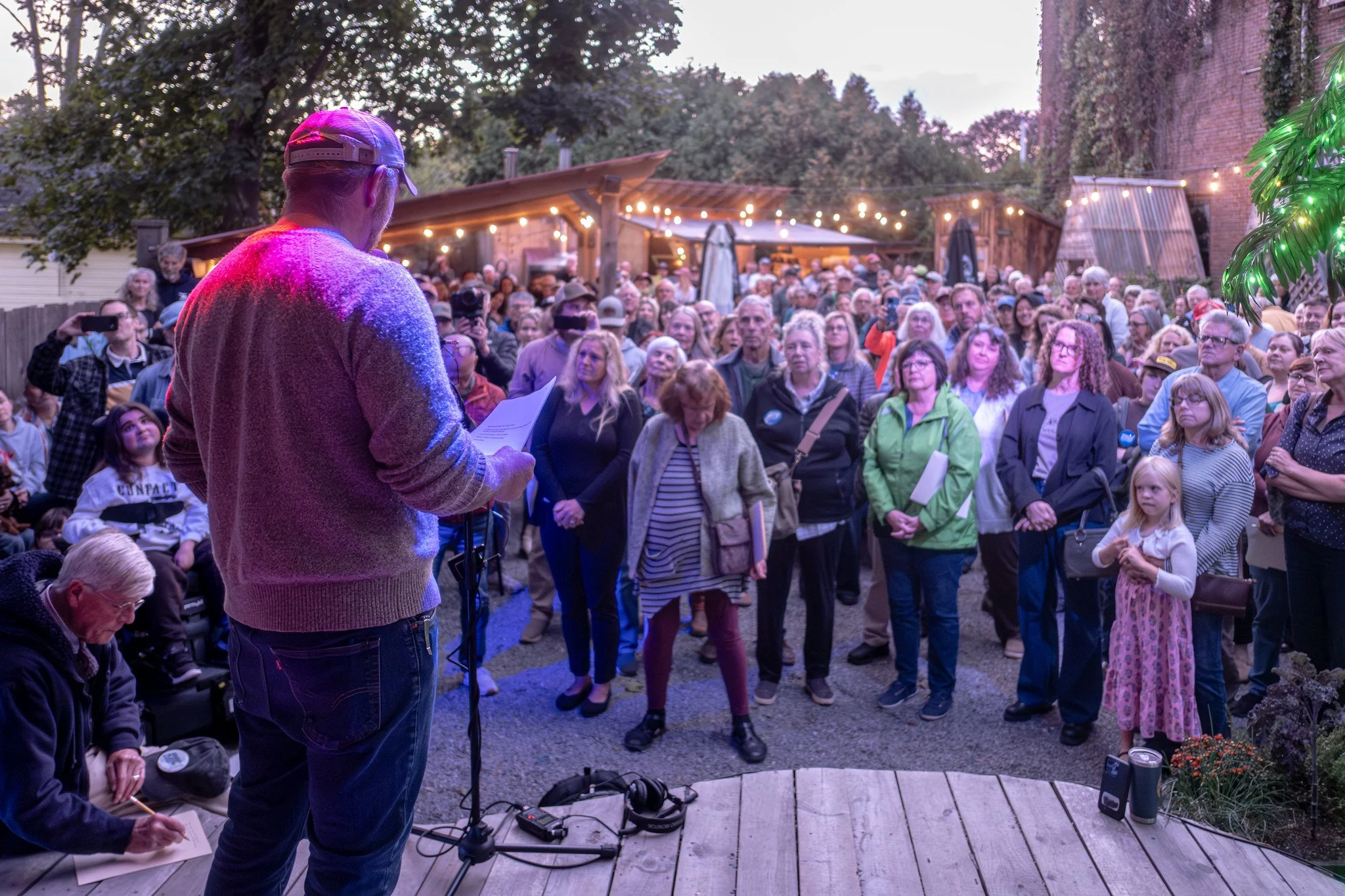 Graham Platner is reading papers on a small stage. He is facing a large outdoor audience during early evening, with trees, string lights, and a wooden building in the background. Photo by Nick Gervin.