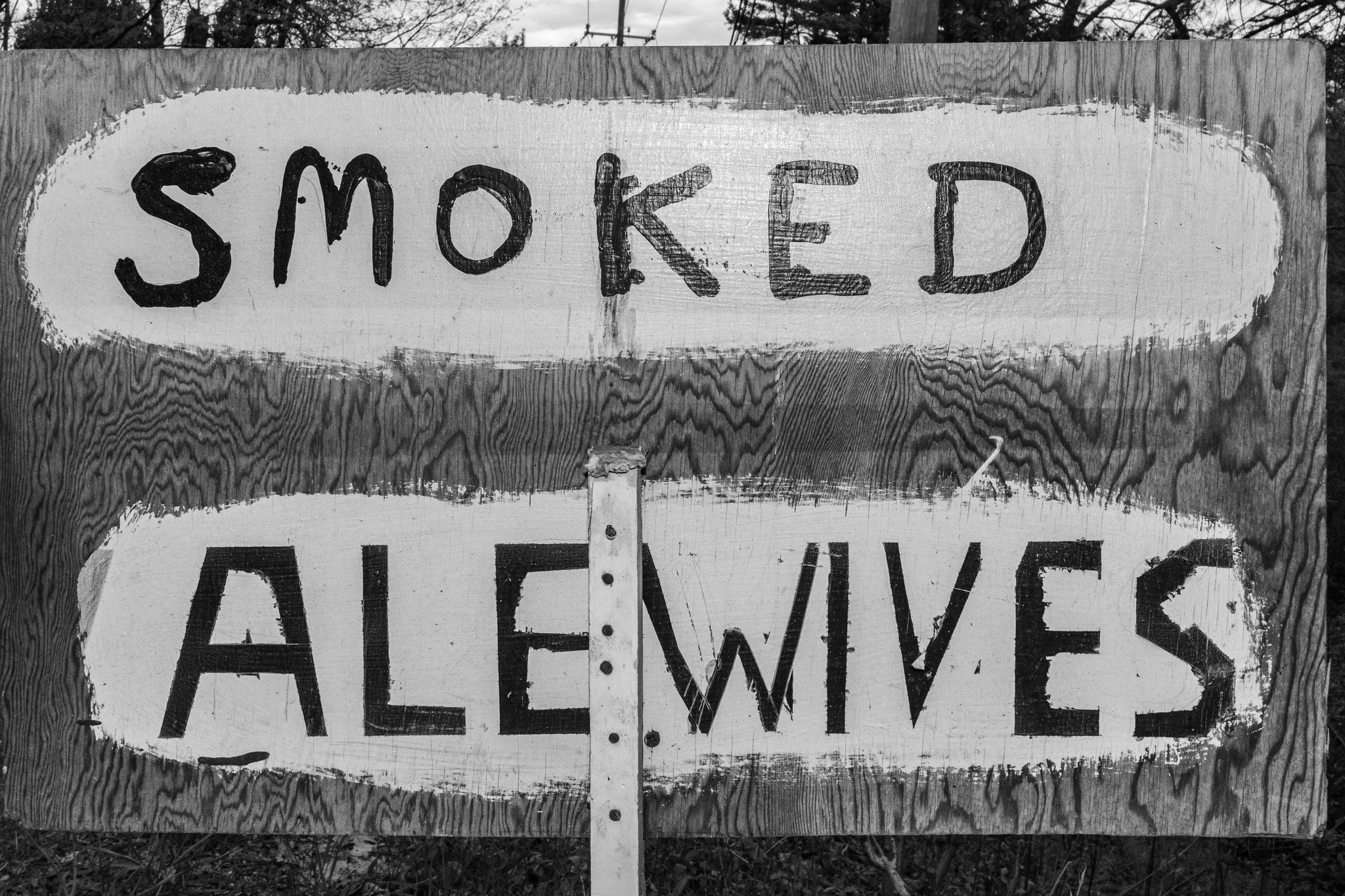 A black and white photo of a wooden sign with the word 'SMOKED' written in bold, black letters inside an oval shape, mounted on a post, with trees in the background.