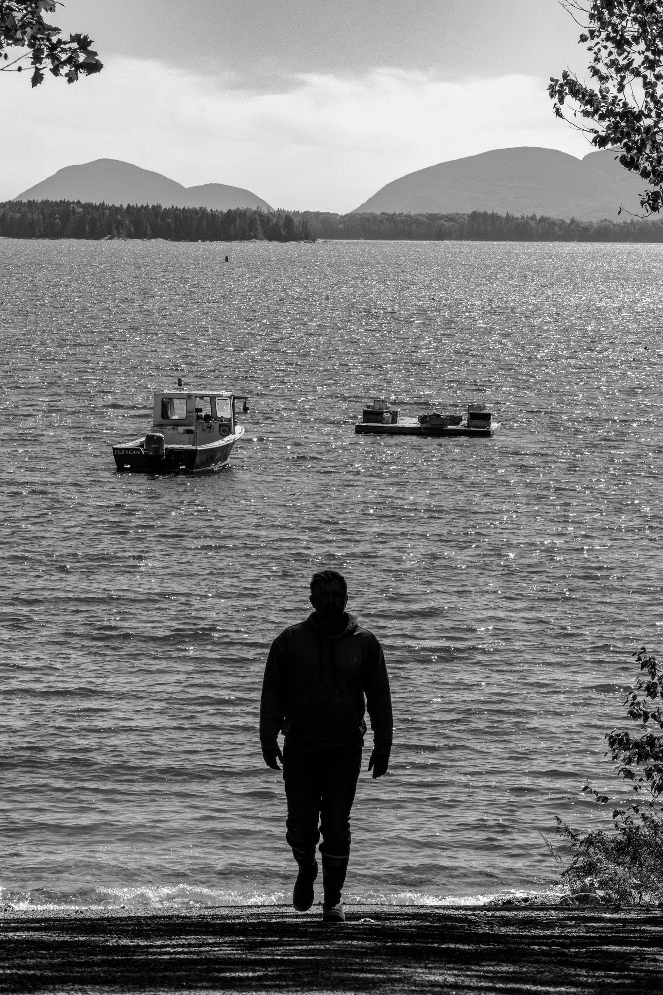 Graham Platner walking along the edge of a body of water with mountains in the distance and boat on the water, black and white photo by Nick Gervin.