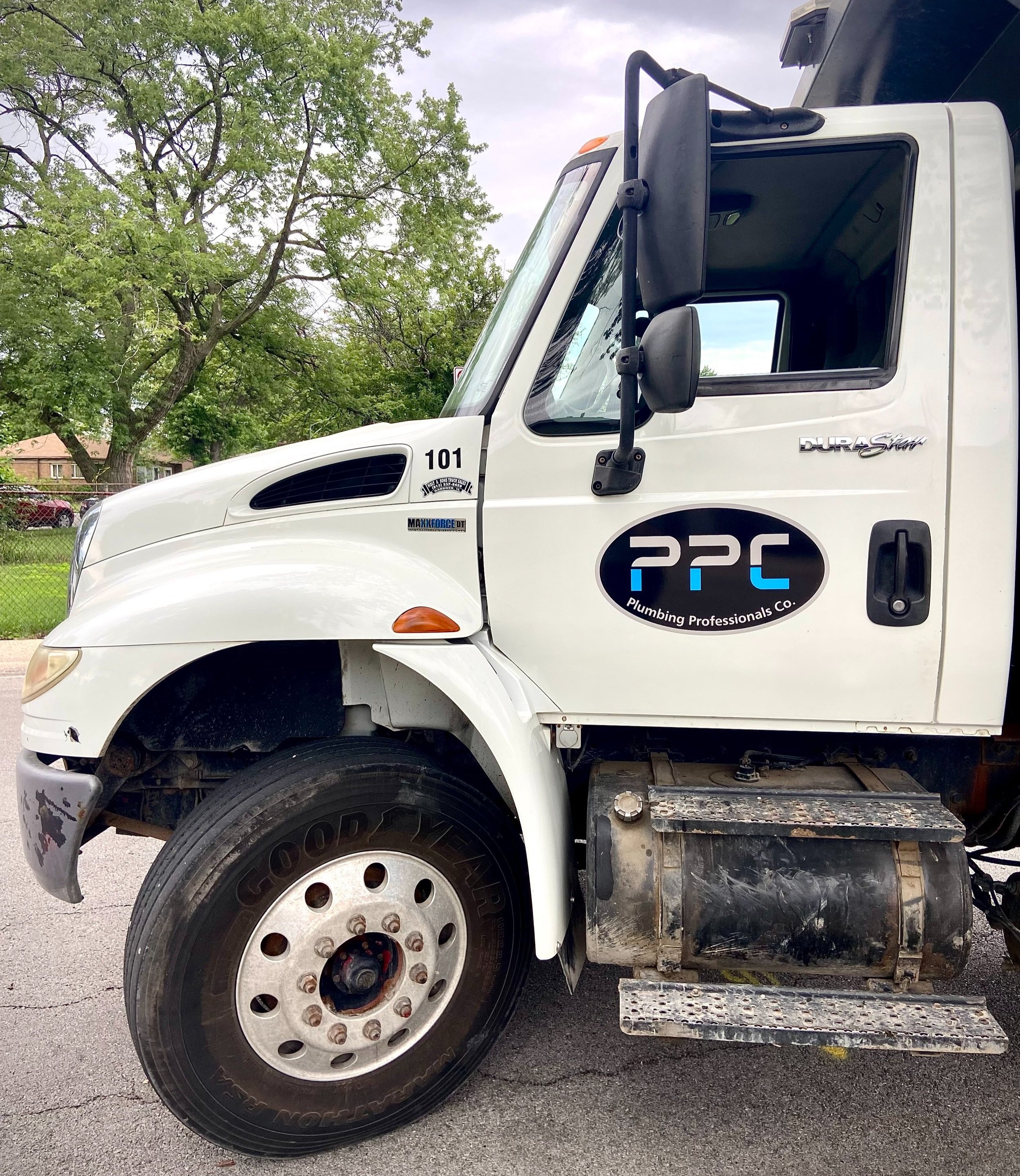 Close-up of a white plumbing truck, showing the front wheel, side mirror, and part of the company's logo. The truck is parked outdoors with green trees and a neighborhood in the background.
