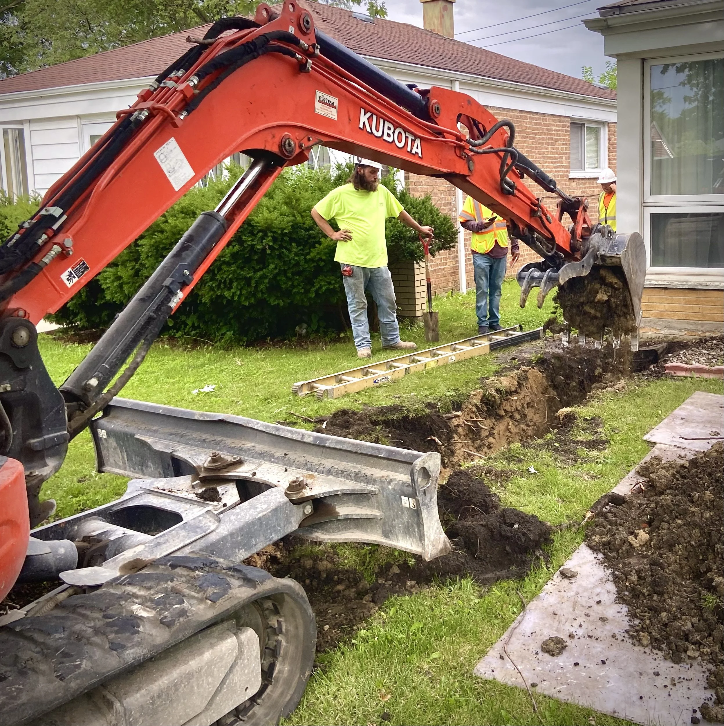 Backhoe excavator digging a trench in a residential yard, with workers supervising, near a house with a brick wall and a window.