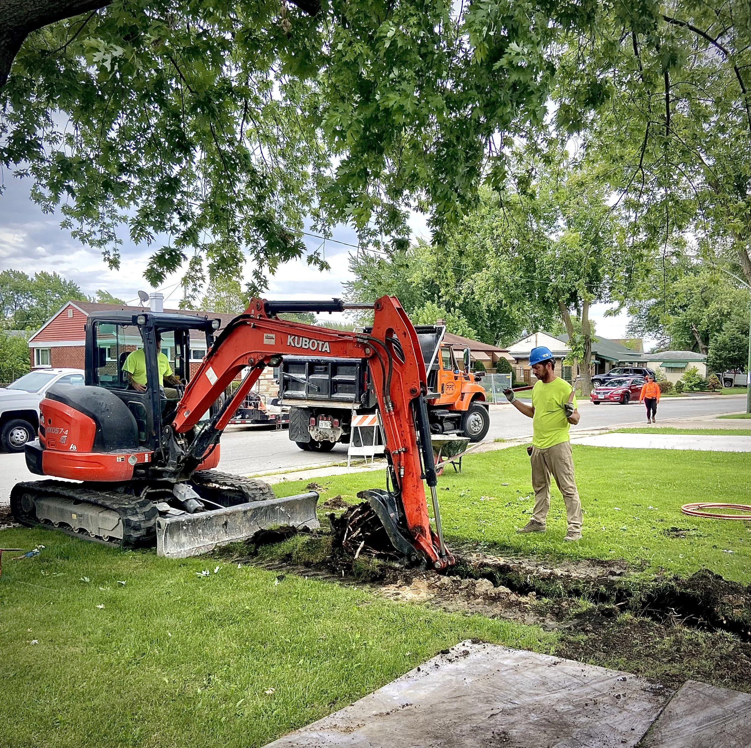 Workers repairing a sidewalk with a small excavator in a residential neighborhood, with houses, trees, and parked cars in the background.