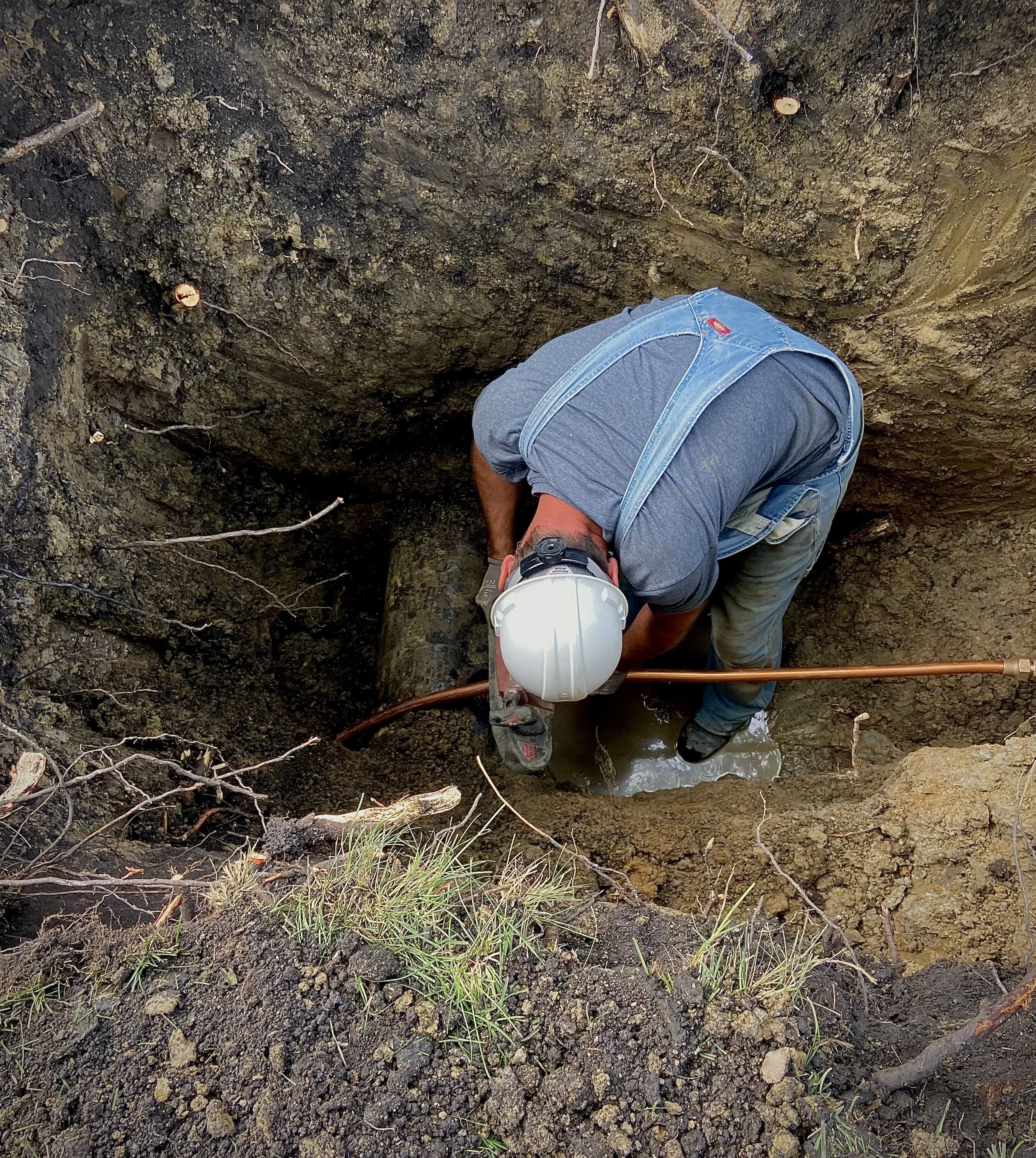 A person wearing a gray t-shirt and jeans with suspenders is working in a deep hole filled with water and muddy soil, using a tool to extract water from the ground.
