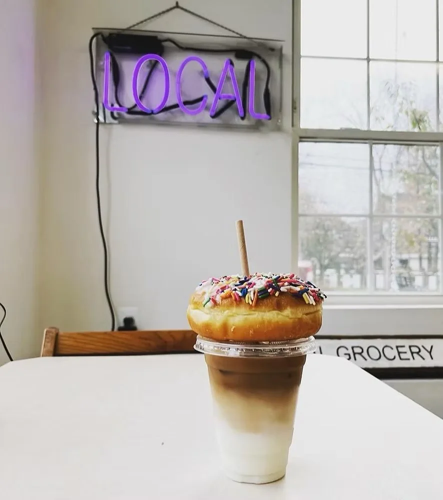 A coffee cup with layered milk and coffee, topped with a glazed donut decorated with colorful sprinkles and a small stick, inside a cafe with a purple neon 'LOCAL' sign and a window in the background.