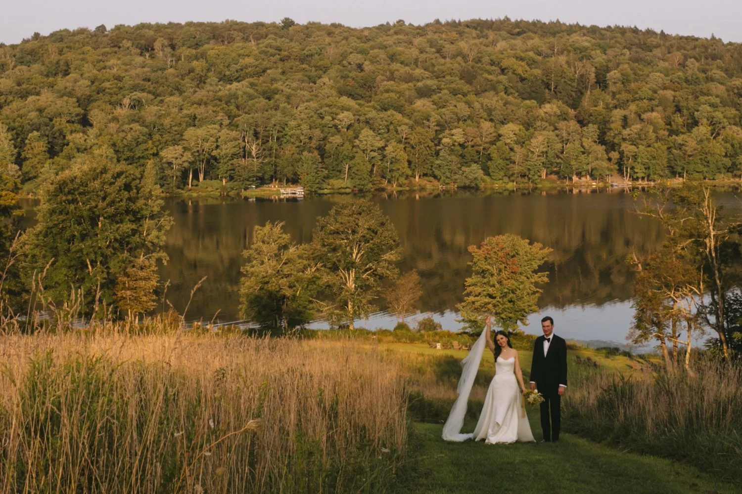 Bride and groom standing on a grassy path by a lake surrounded by trees and hills, dressed in wedding attire, during sunset.