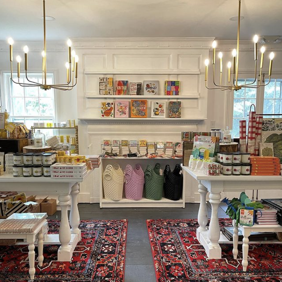 View of a boutique store with white furniture, colorful bags, stationery, and books displayed on shelves and tables, with two matching chandeliers hanging from the ceiling and patterned rugs on the floor.