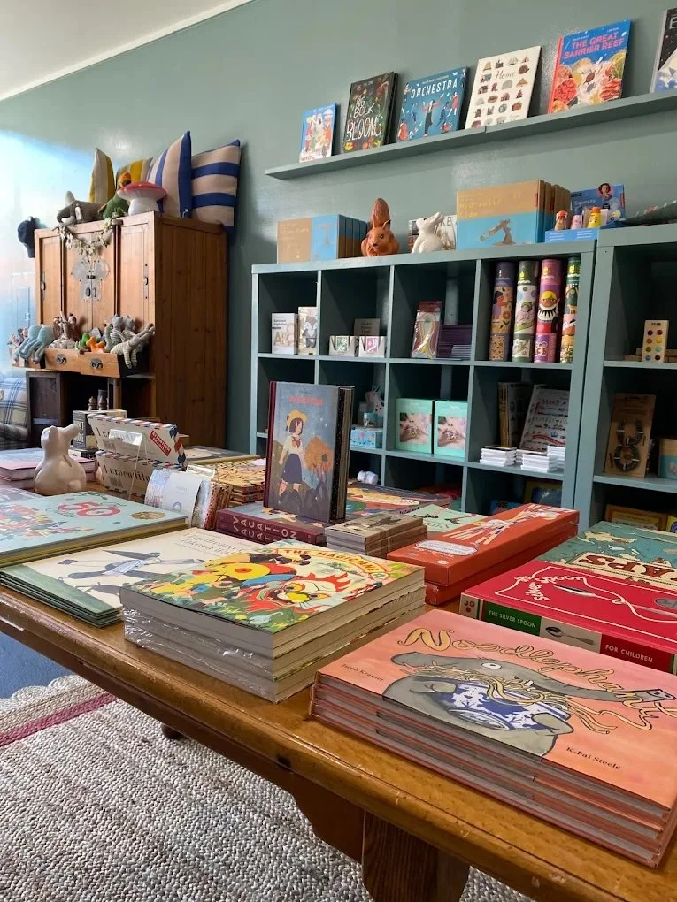 Display of children's books, toys, and stuffed animals on a table and shelves in a cozy shop or library.
