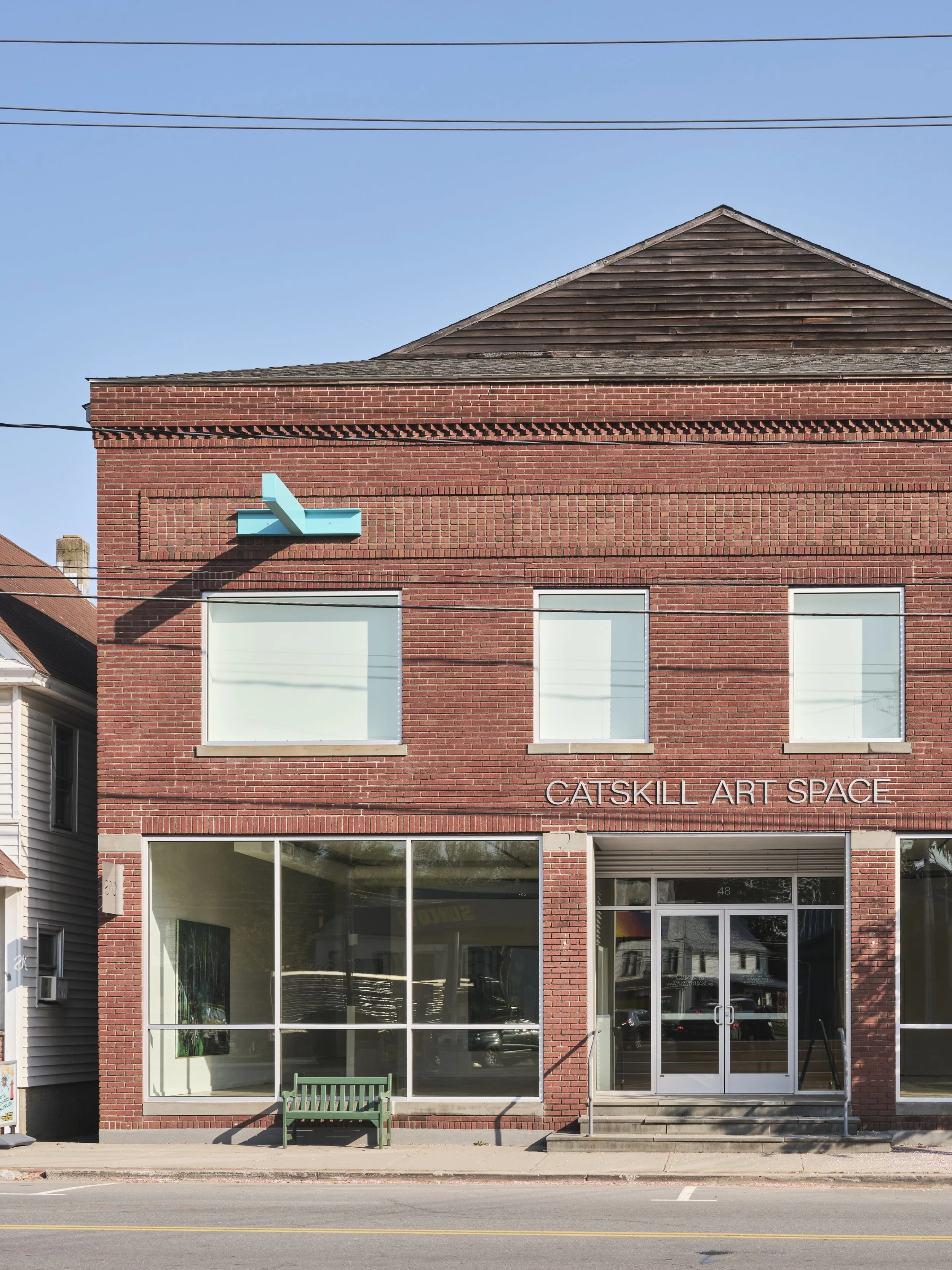 Front view of a red brick building with large glass windows and a sign that reads 'Catskill Art Space'. There is a small green bench outside and pastel blue abstract art installation on the upper left corner of the building.